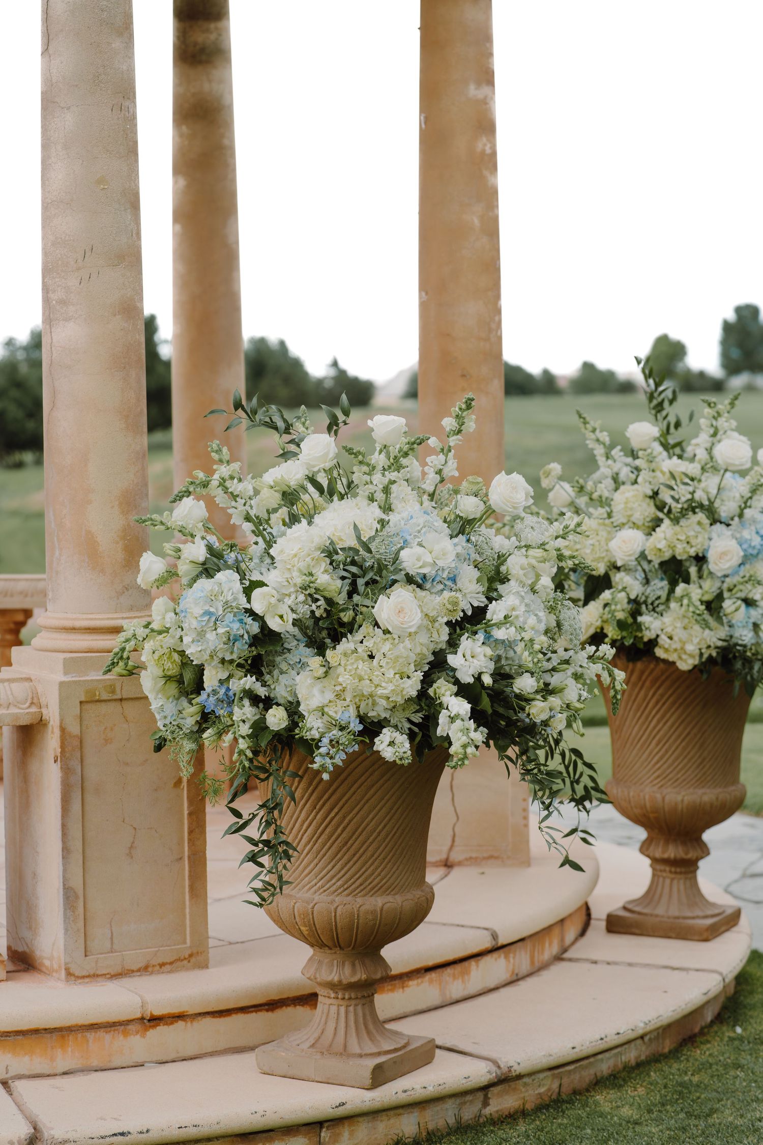 Elegant stone urns filled with white and blue flowers line a curved outdoor colonnade for a wedding ceremony.