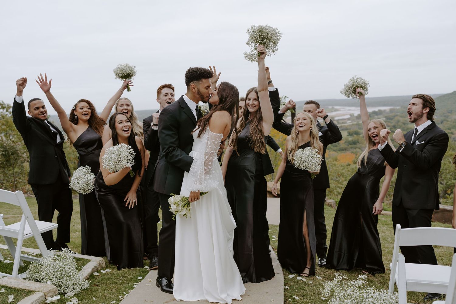 Wedding celebration with bridal party in black attire cheering outdoors against scenic backdrop.