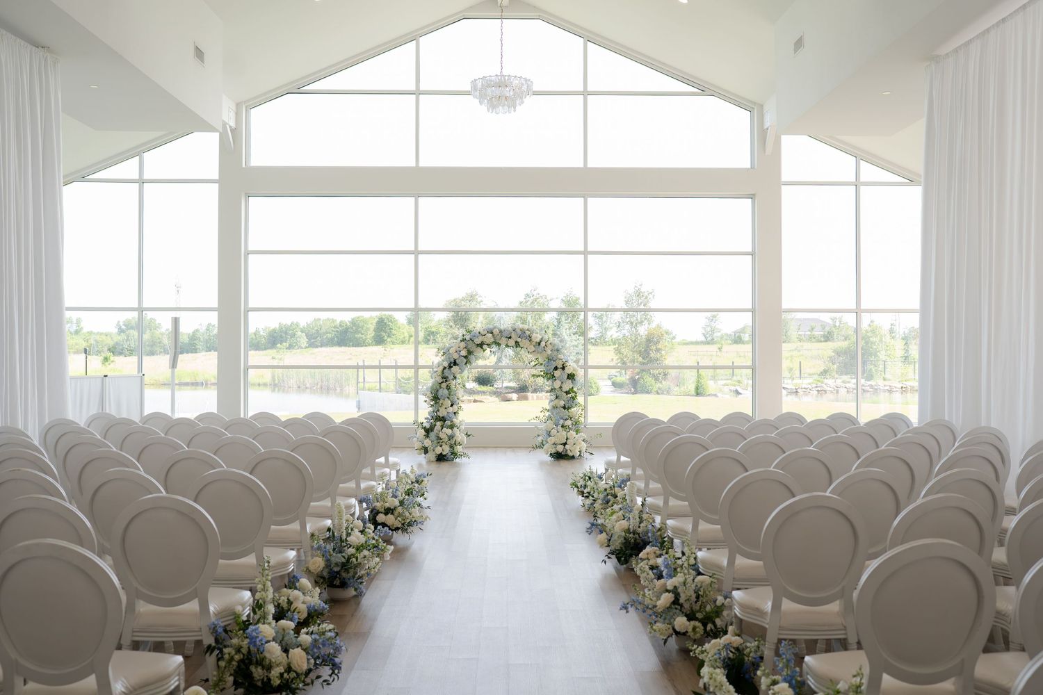 Elegant white wedding ceremony venue with floral arch, draped walls, and blue flower arrangements lining the aisle.
