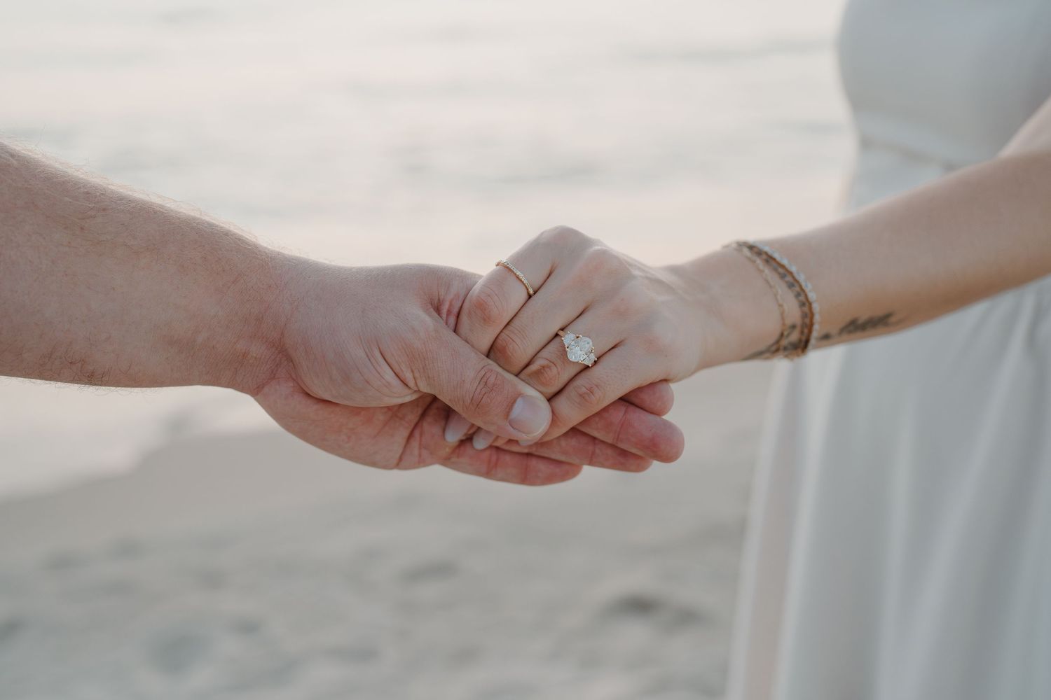 Close-up of hands displaying an engagement ring against beach backdrop.