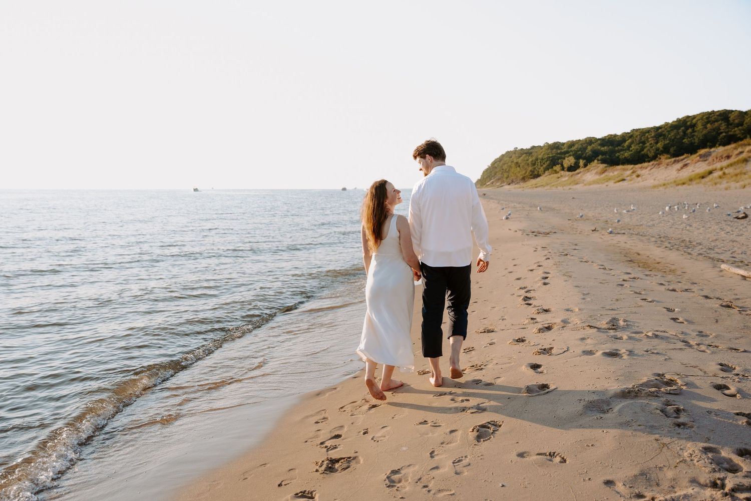 A romantic beach photoshoot showing a couple walking barefoot on the sand at sunset.