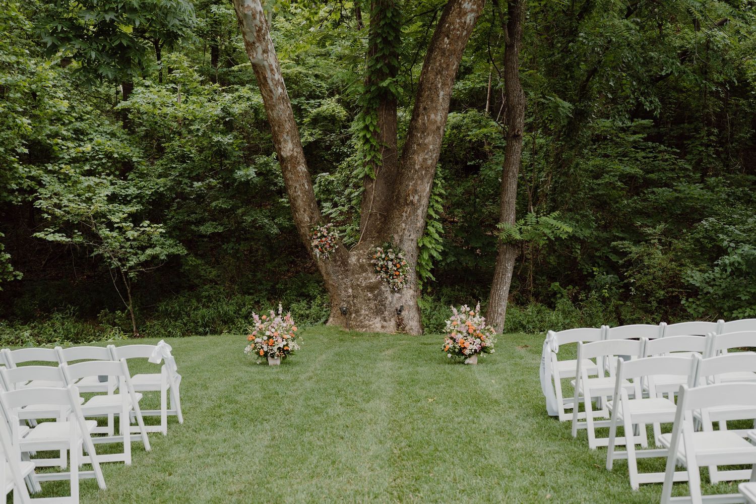 White ceremony chairs arranged in rows on a lawn surrounded by trees.