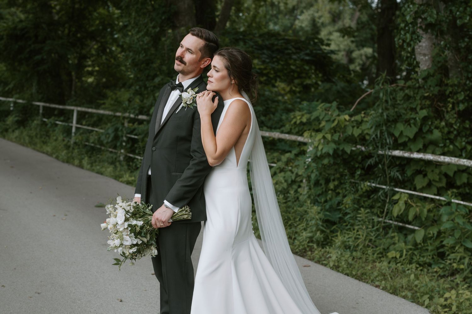 A romantic wedding couple poses on a tree-lined path with the bride holding a white floral bouquet.
