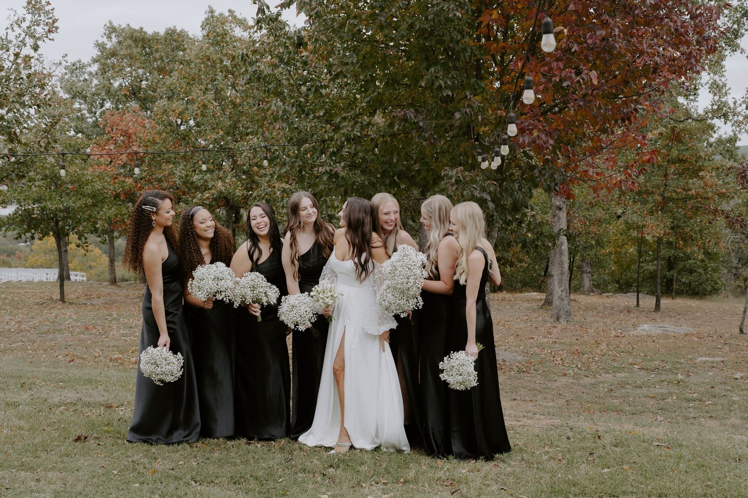 A bridal party in black dresses poses together on grass with autumn trees in the background.
