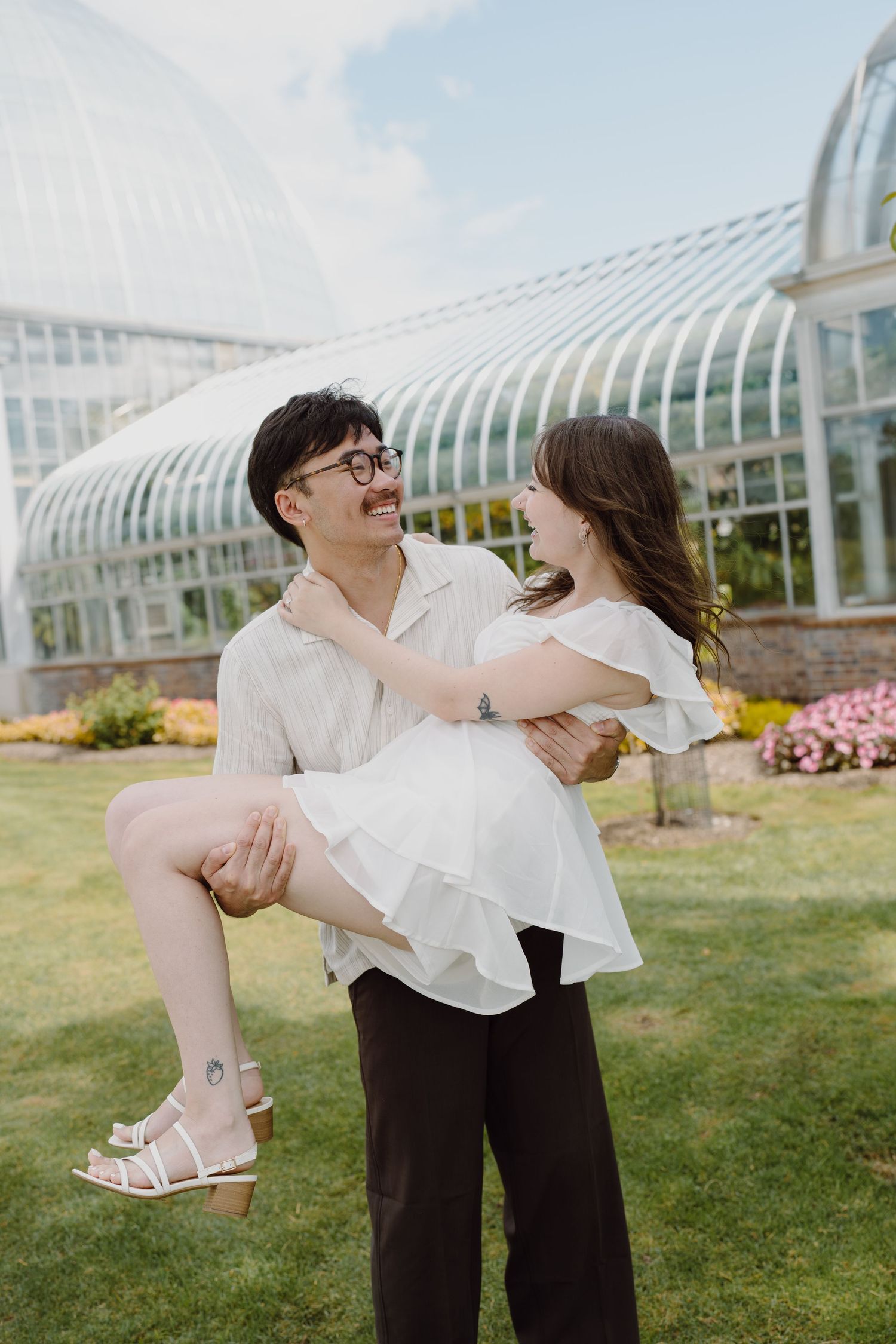 Dancing figures embrace in front of a greenhouse with white architectural details and flowering gardens.