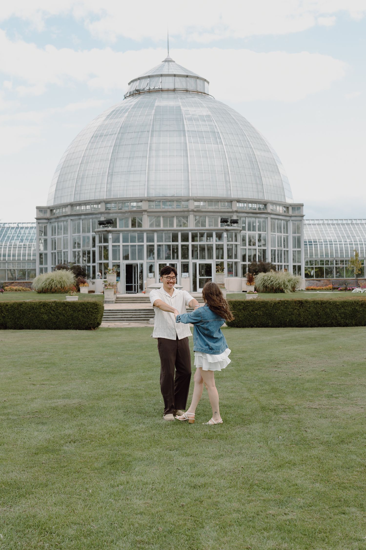 Two people stand on a manicured lawn with a grand glass-domed botanical garden building in the background.