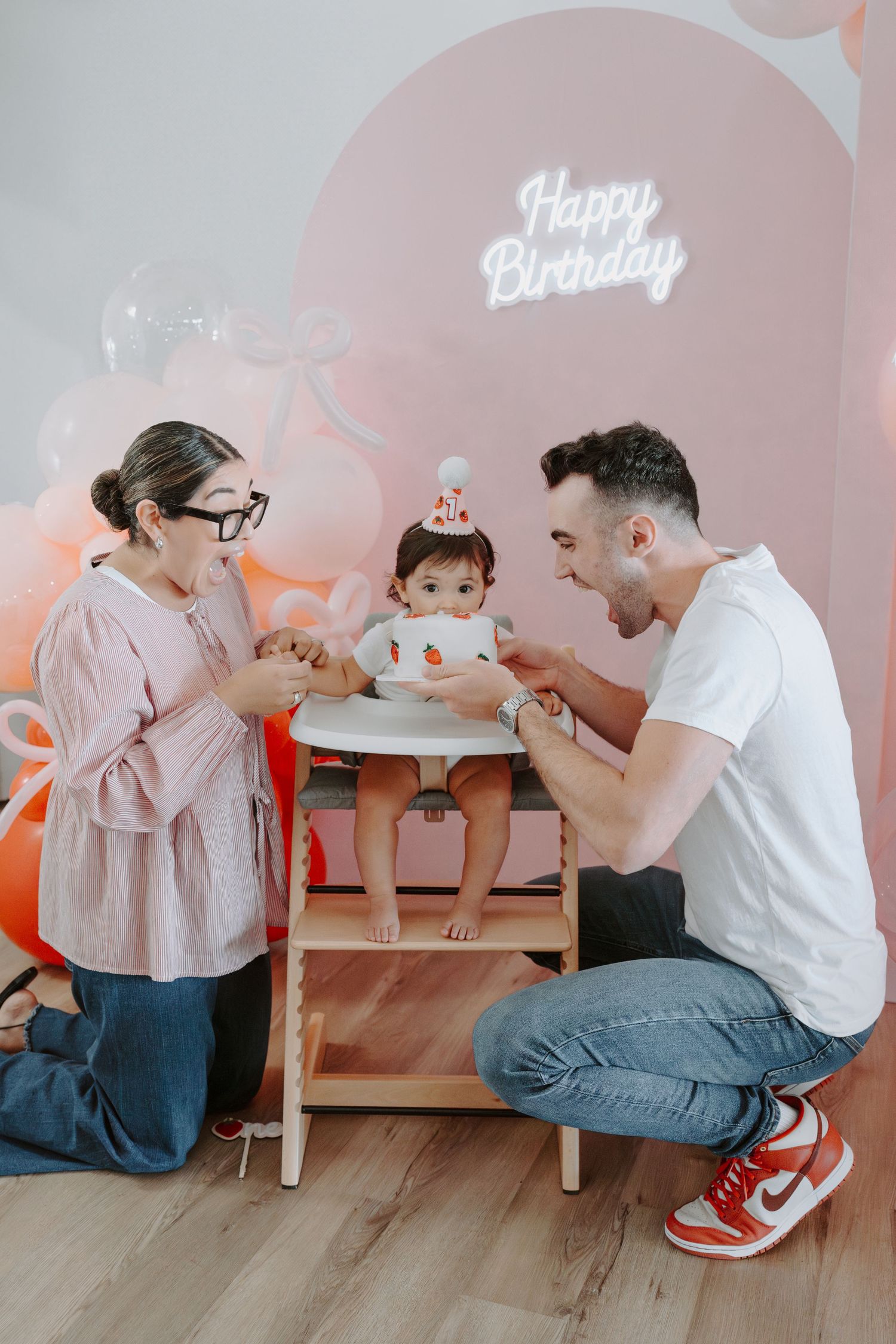 Parents sharing joyful moment with baby during birthday celebration with pink decor.