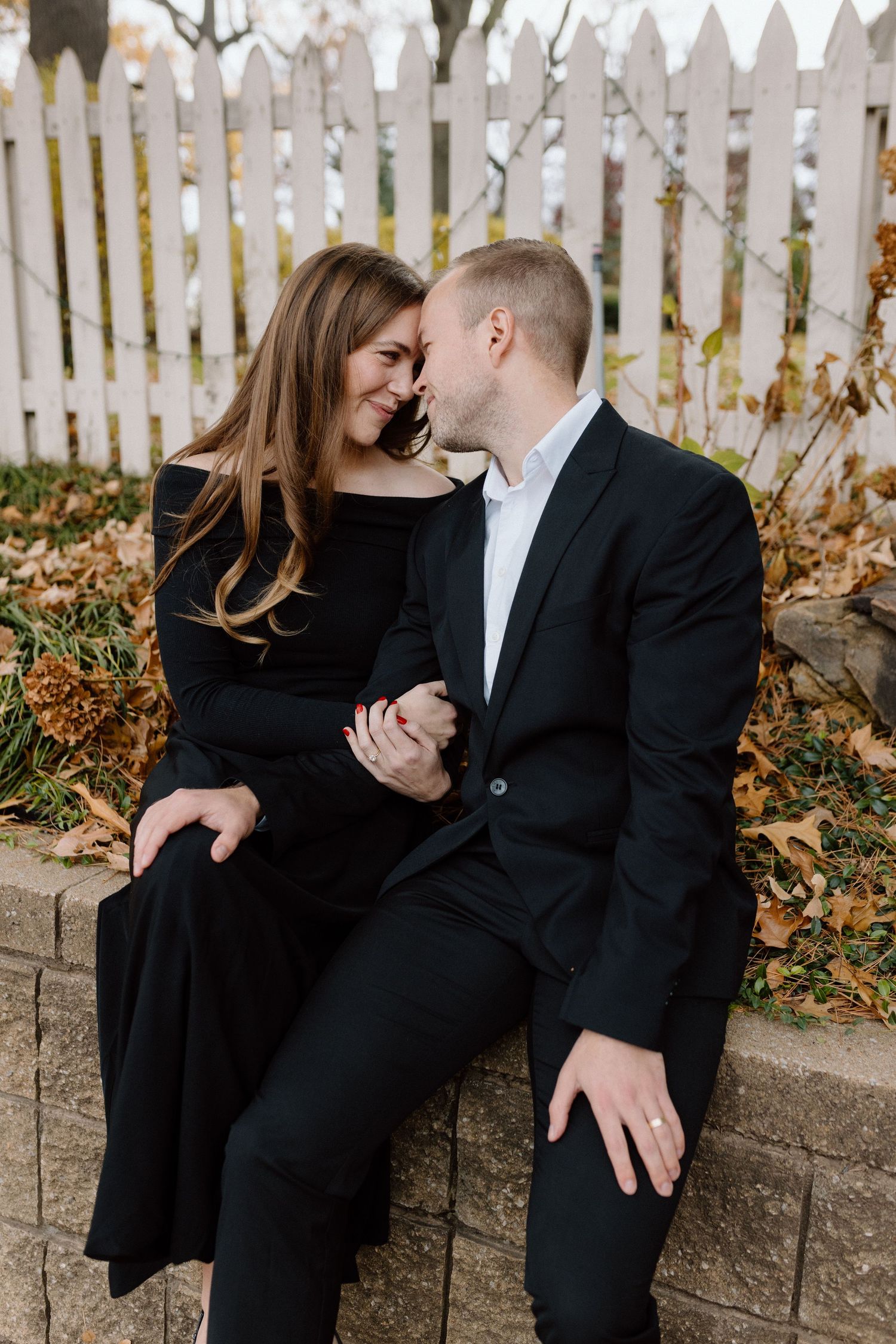 Couple in formal black attire share romantic moment by white picket fence during fall engagement session.