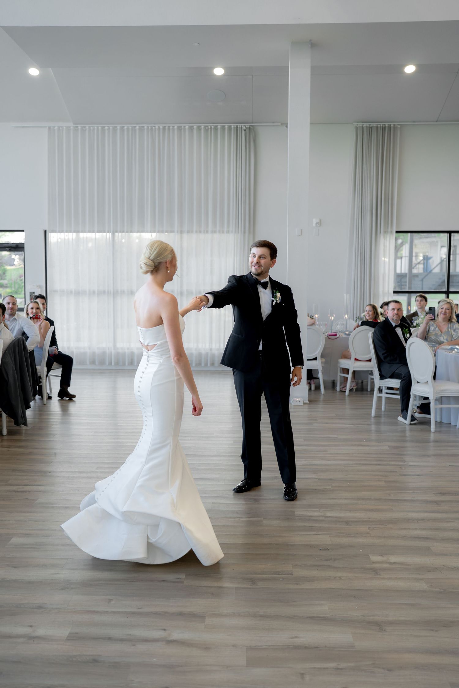 Bride and groom share their first dance in an elegant white ballroom while wedding guests look on and applaud.