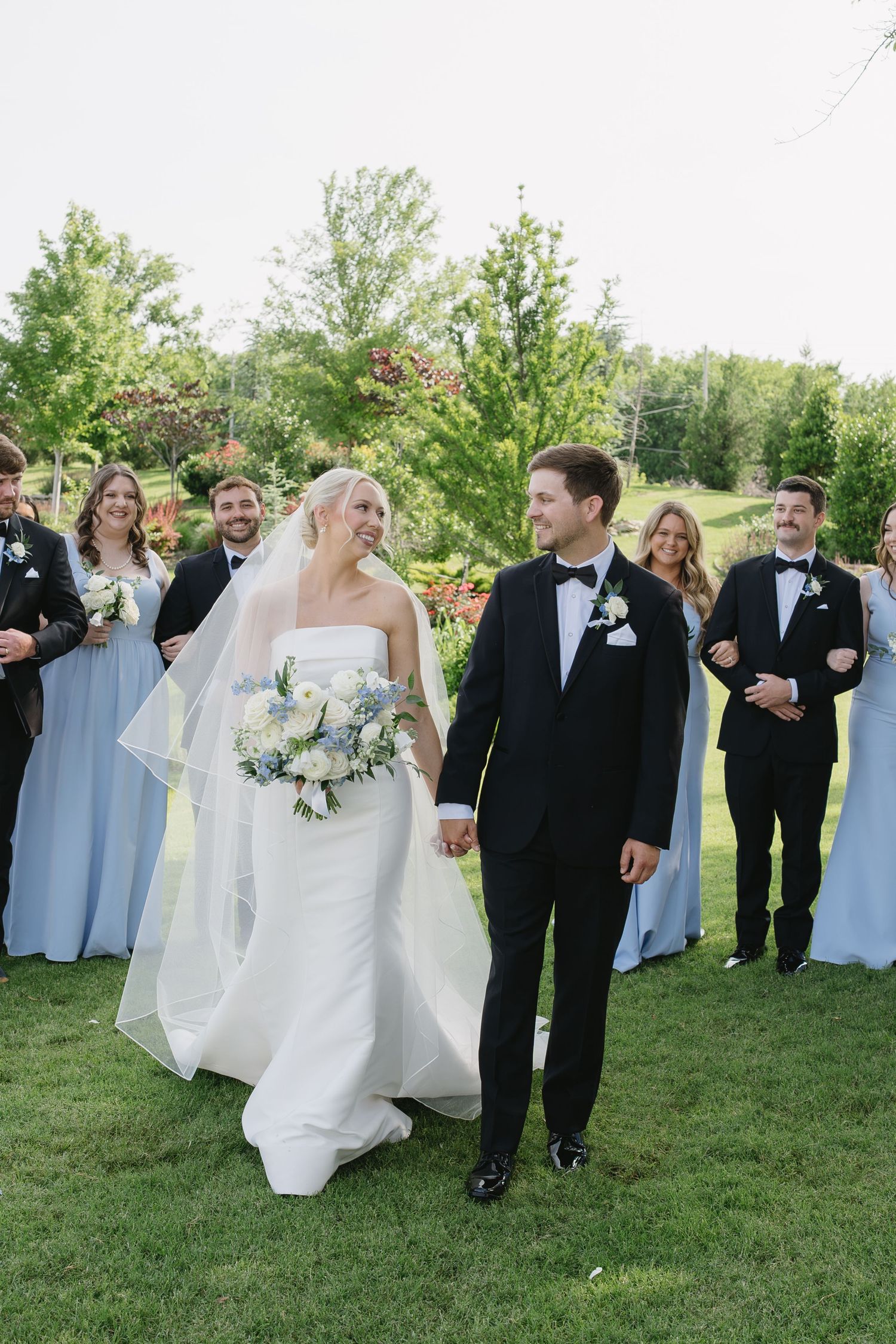 Bride and groom walk hand-in-hand on green lawn with wedding party in blue dresses and black tuxedos behind them.