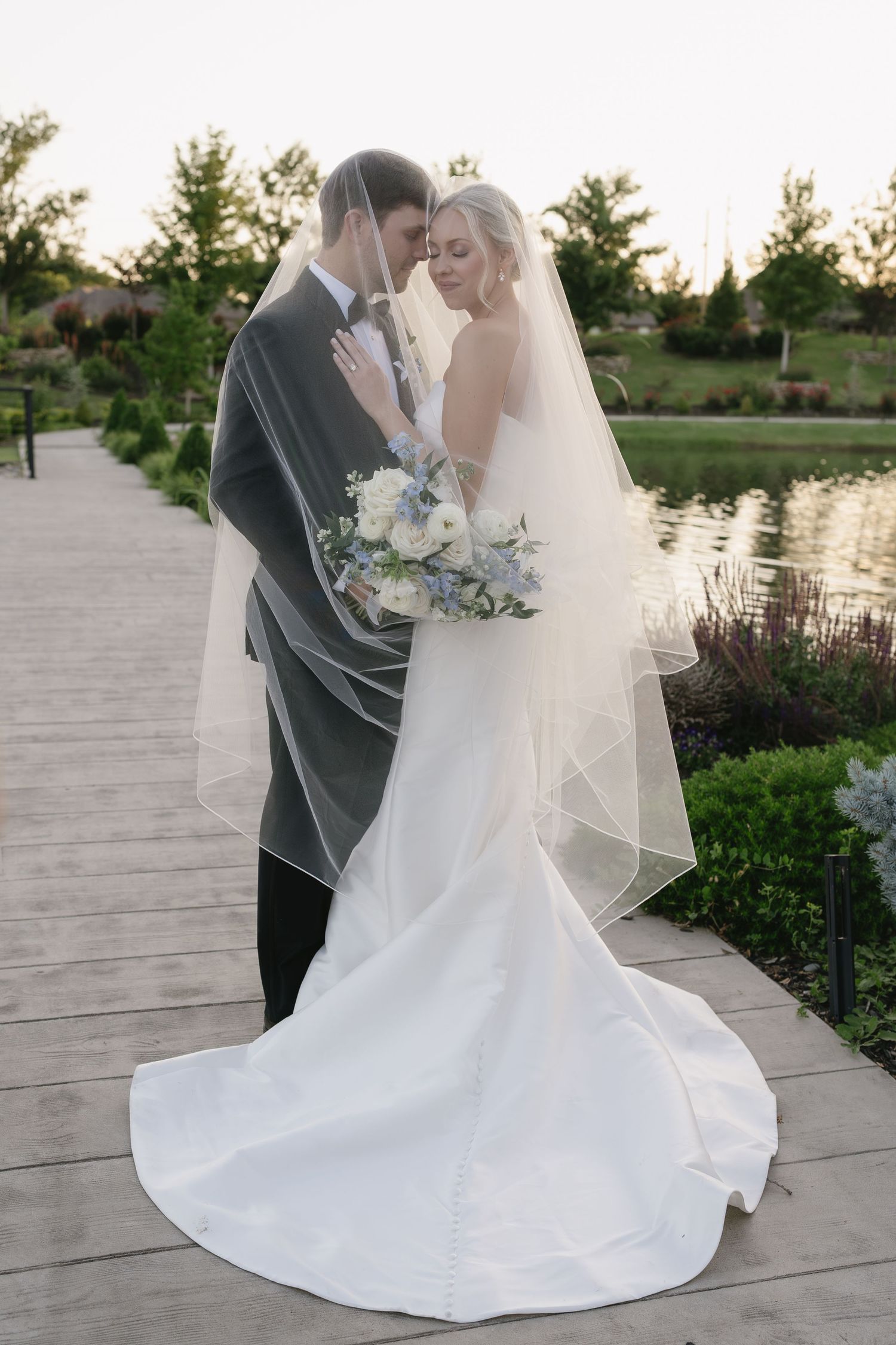 Bride and groom share romantic moment under veil on garden boardwalk, bride holding blue and white bouquet.