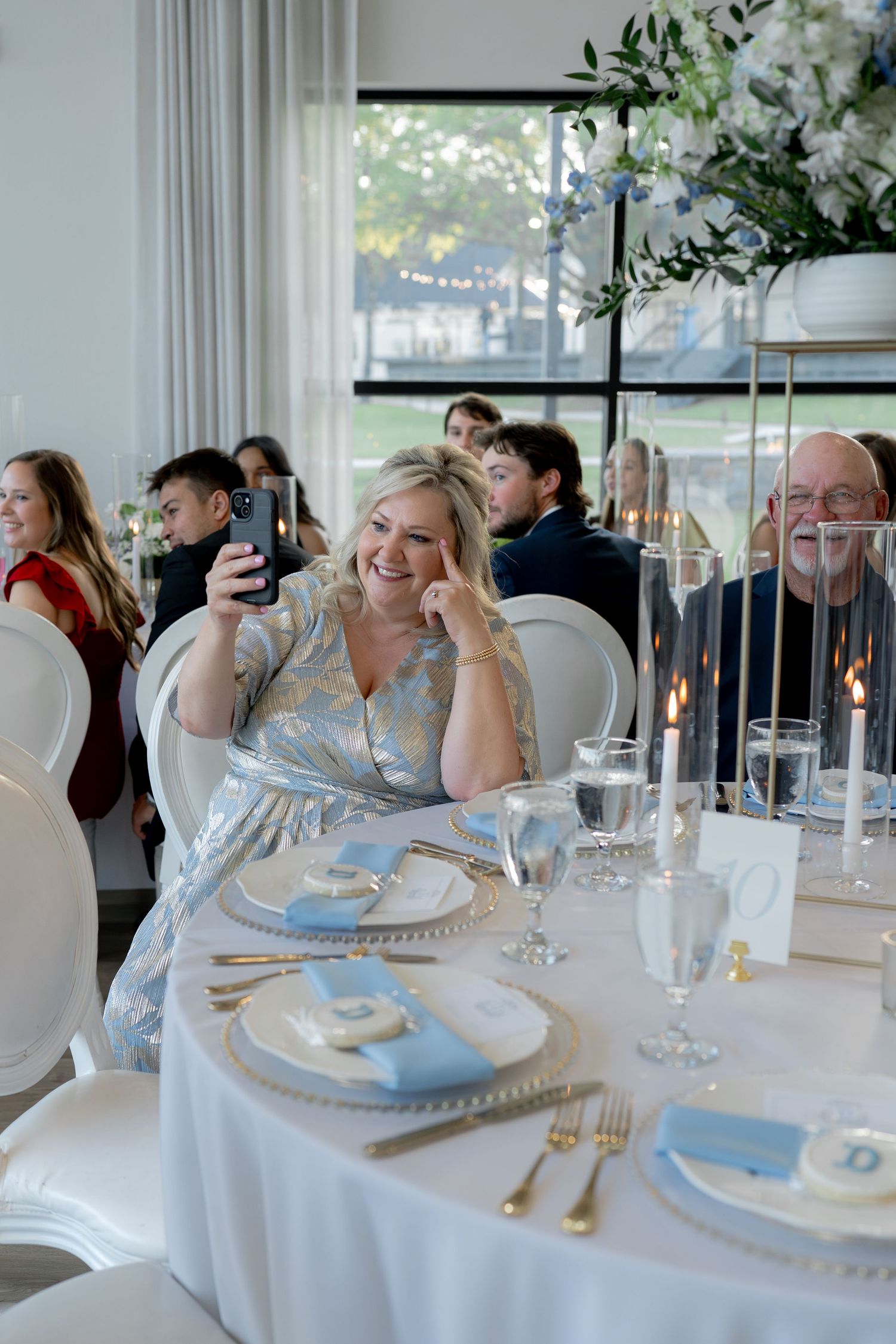 Smiling woman in blue floral dress takes selfie at elegant wedding reception table with gold flatware and candles.