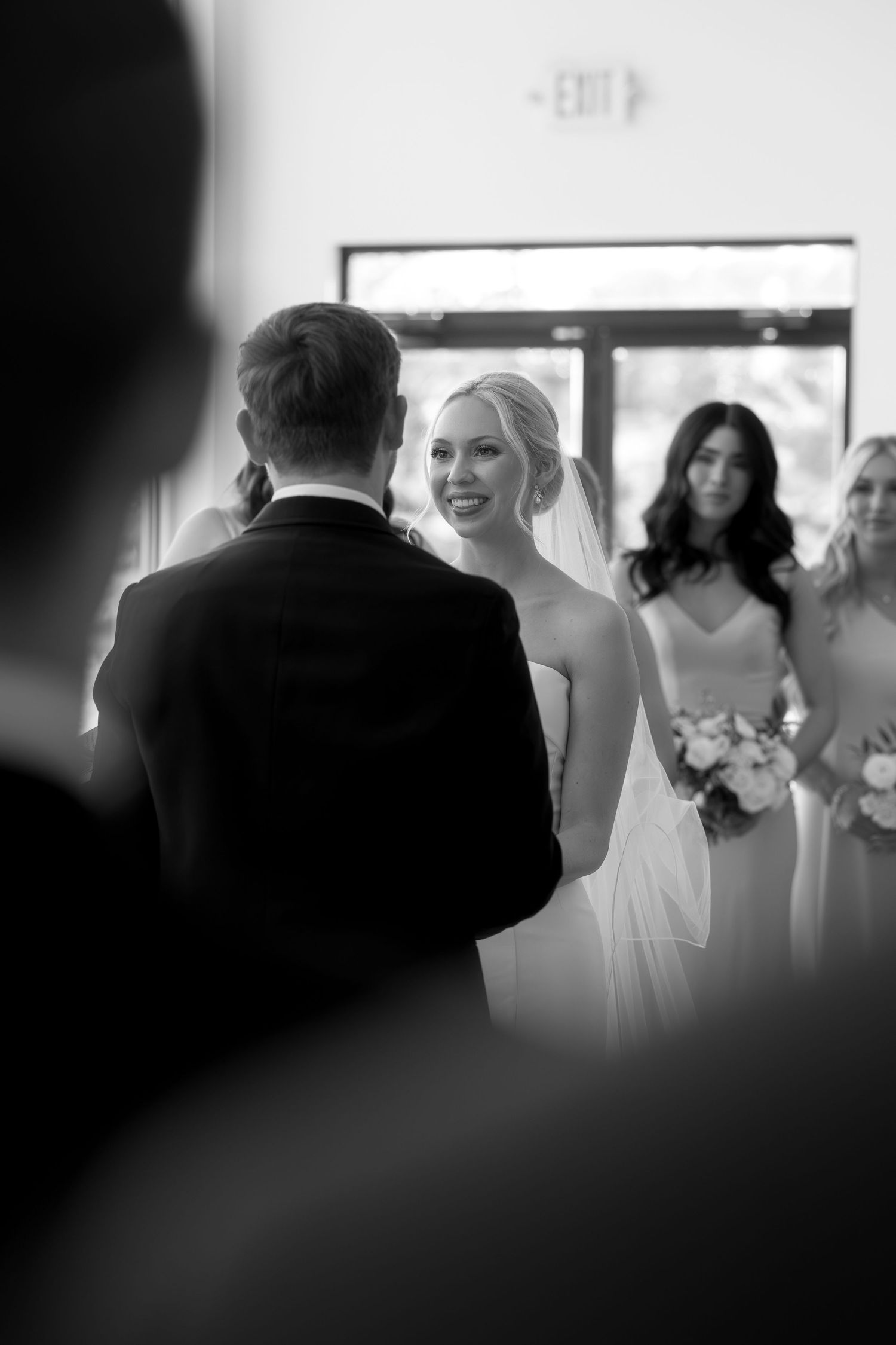 Black and white wedding ceremony photo of smiling bride in veil facing groom, with bridesmaids holding bouquets in background.