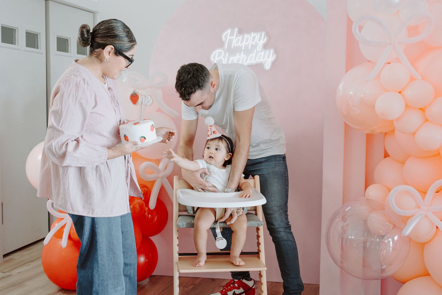 Family celebrating birthday with pink and white balloon decorations and neon signage.