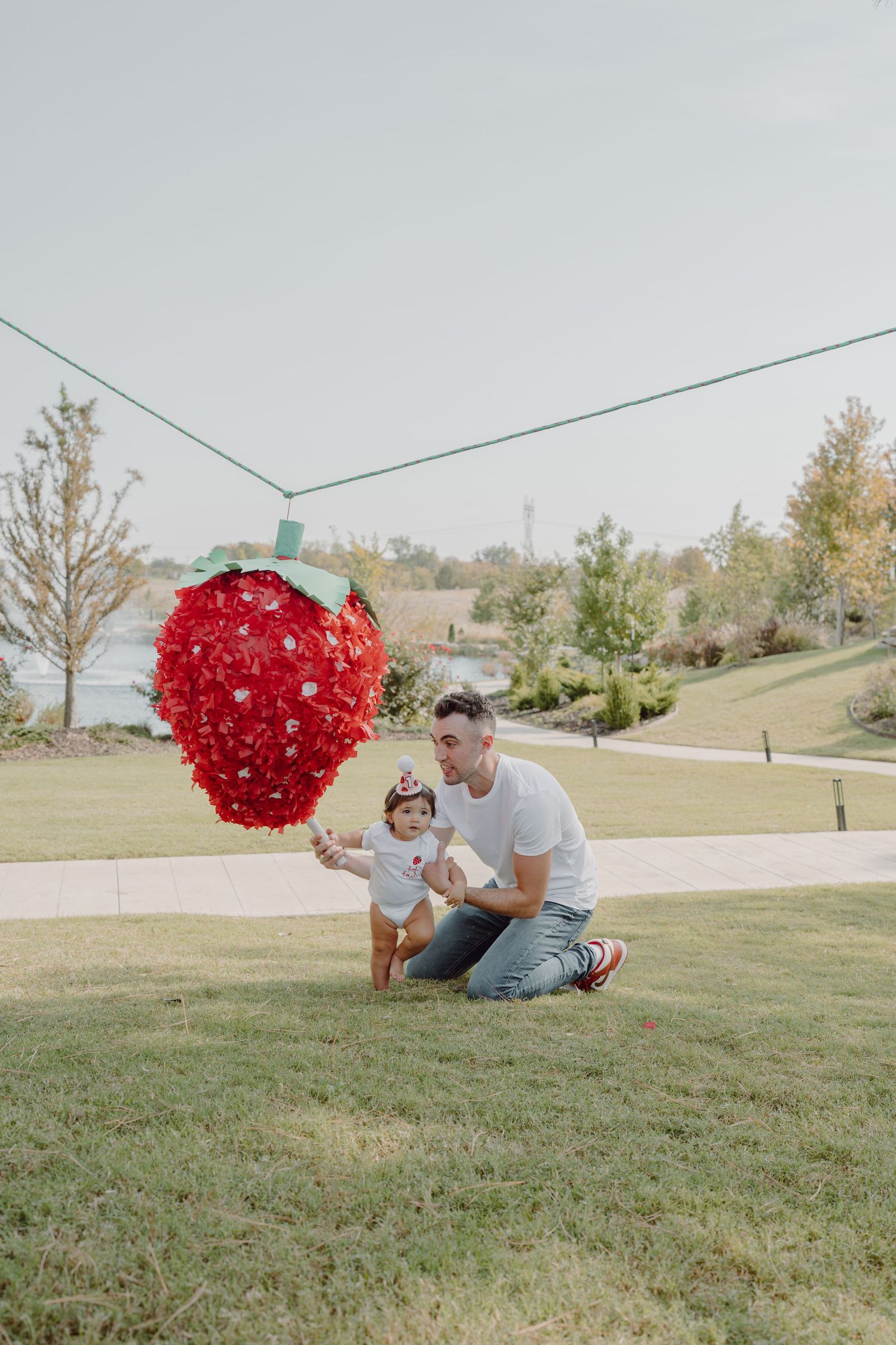 People play with a large red heart decoration on a sloping green lawn near water.