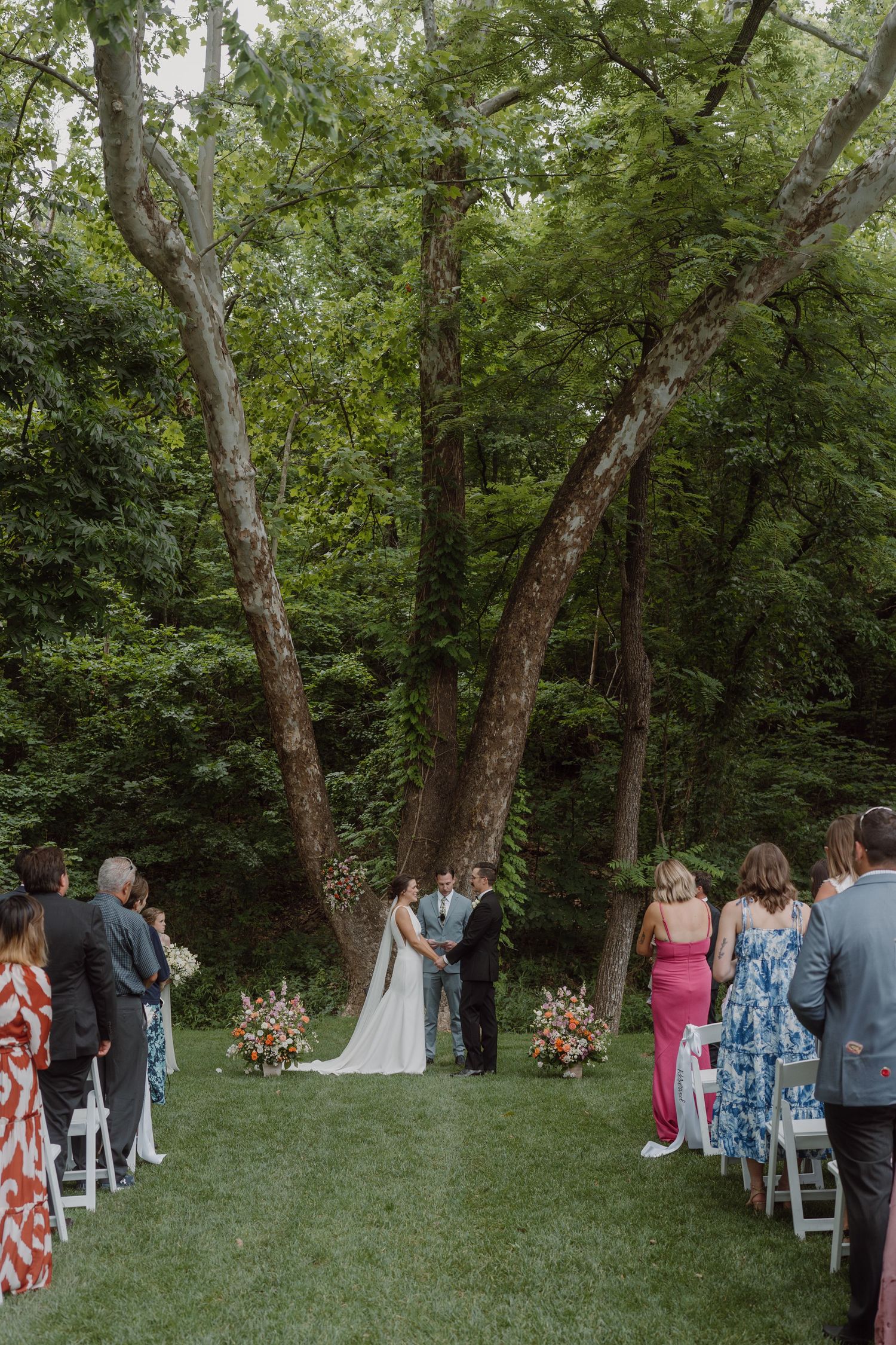 Wedding guests seated in white chairs on a grassy lawn surrounded by trees during an outdoor ceremony.