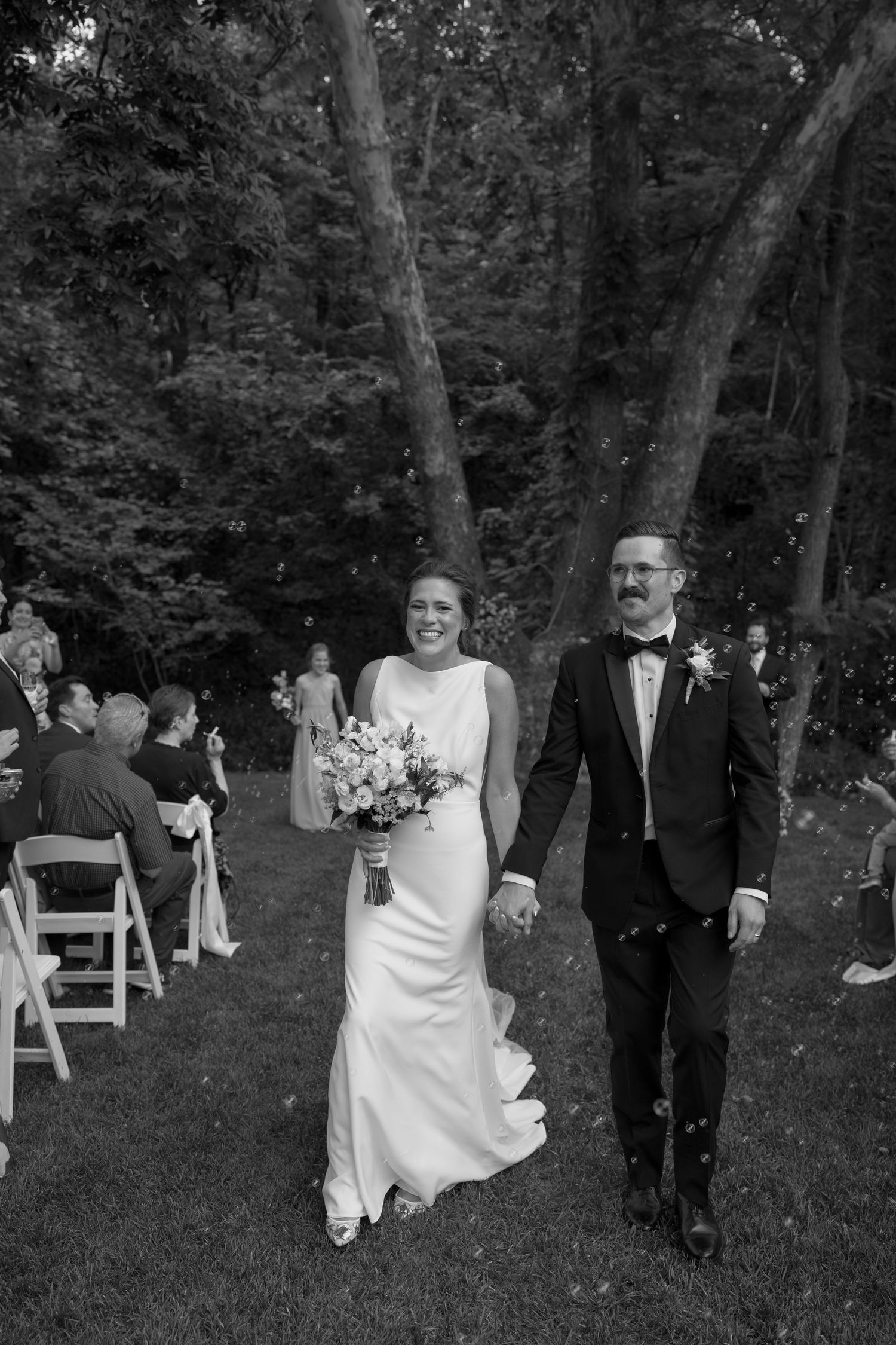 Black and white photo captures a joyful wedding recessional in a wooded outdoor venue.
