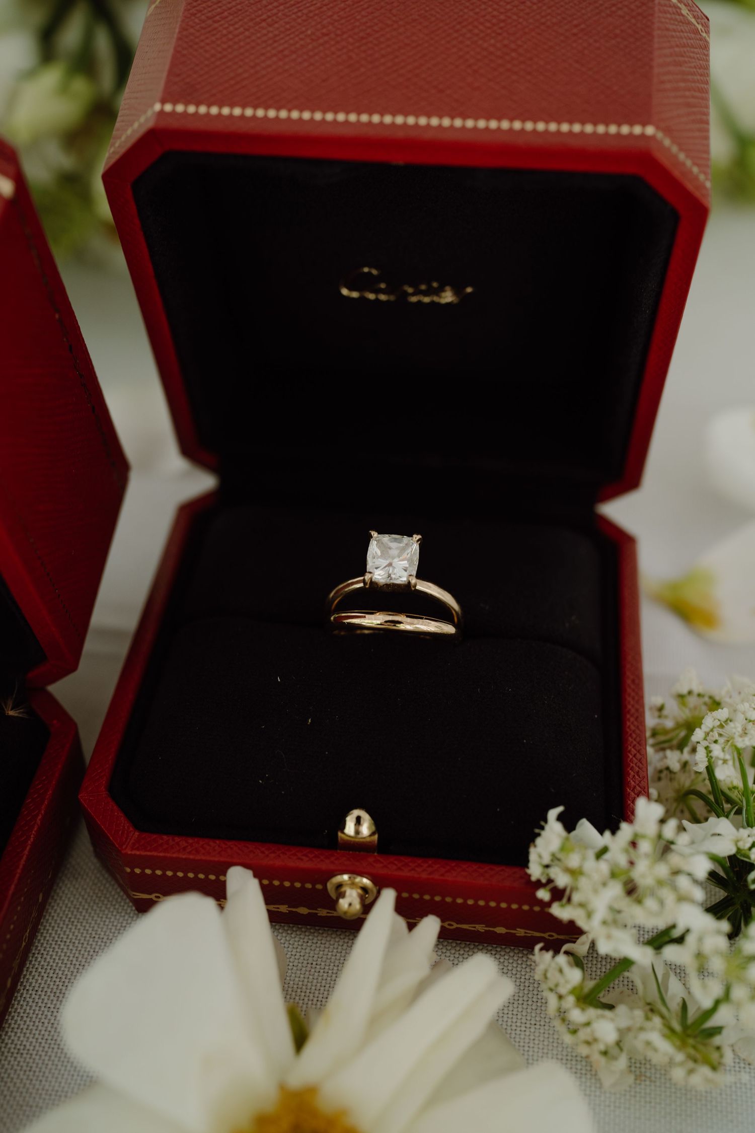 Diamond engagement ring in red velvet box surrounded by white daisies.