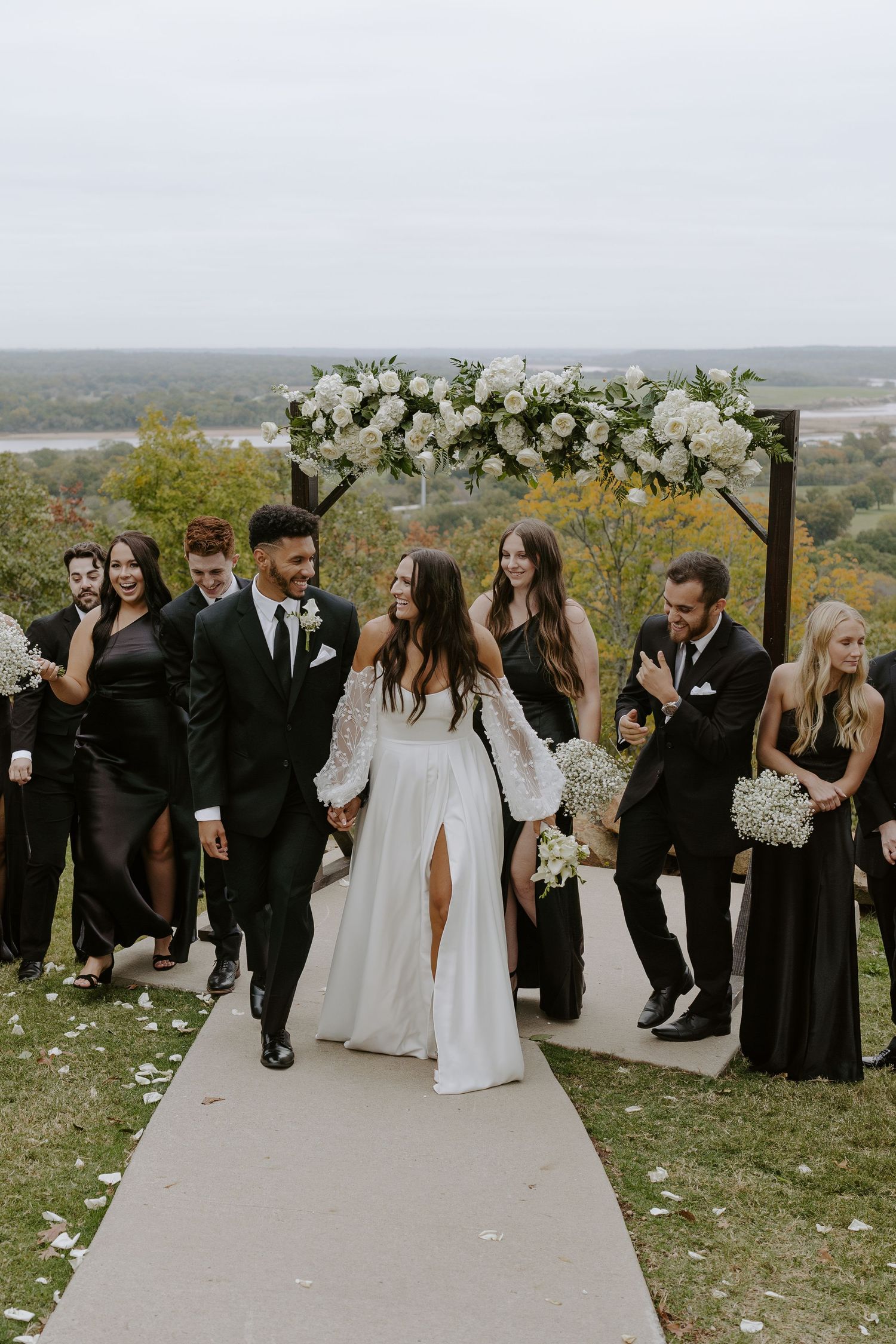 Wedding celebration on a hilltop path with guests in black attire and white floral decorations.