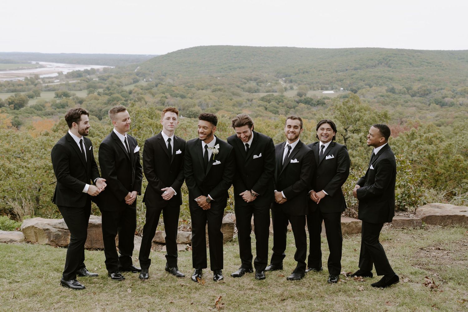 Groomsmen in black suits pose together on a scenic hilltop overlook.
