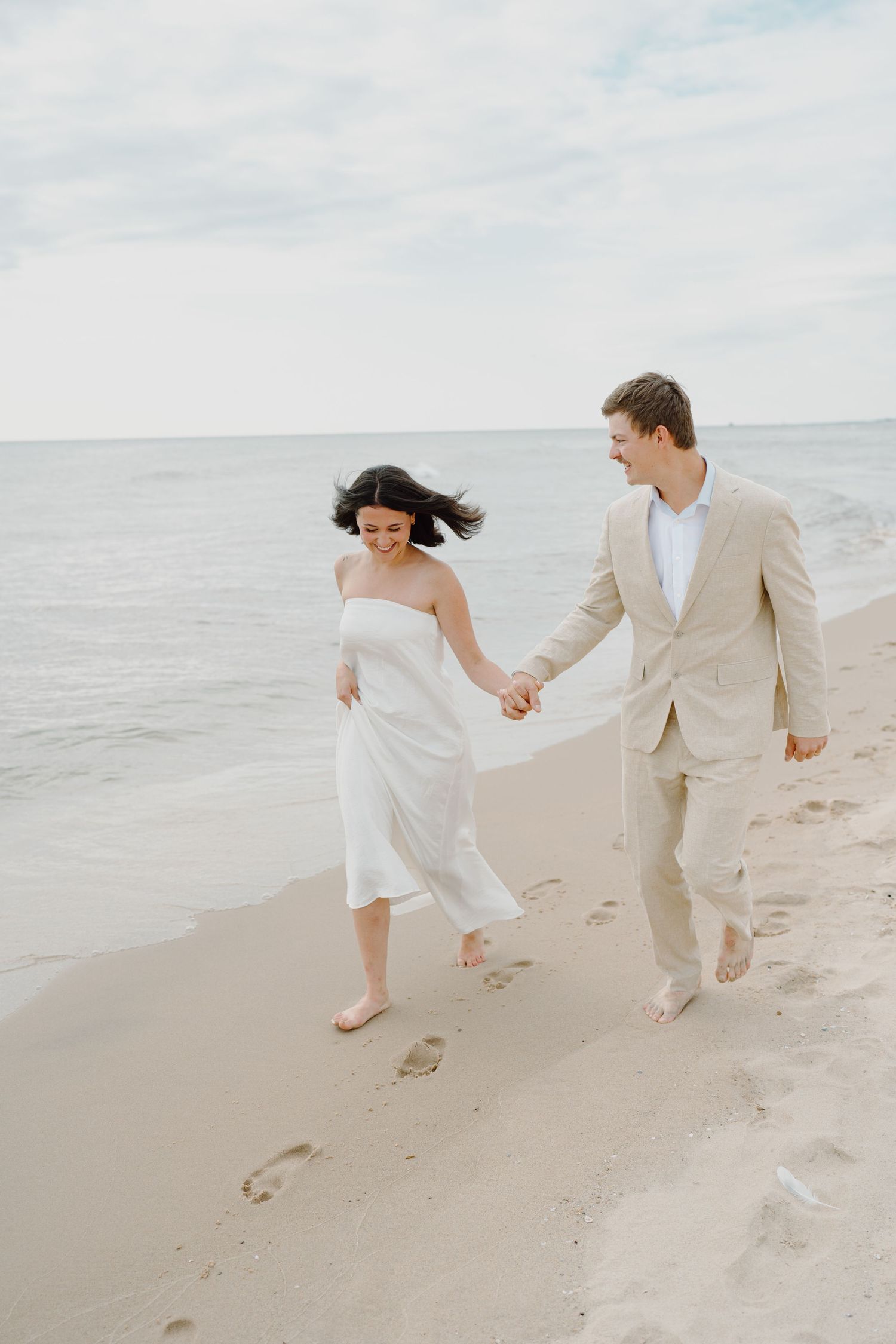 Playful couple running and holding hands along a serene beach shoreline in white and beige outfits.
