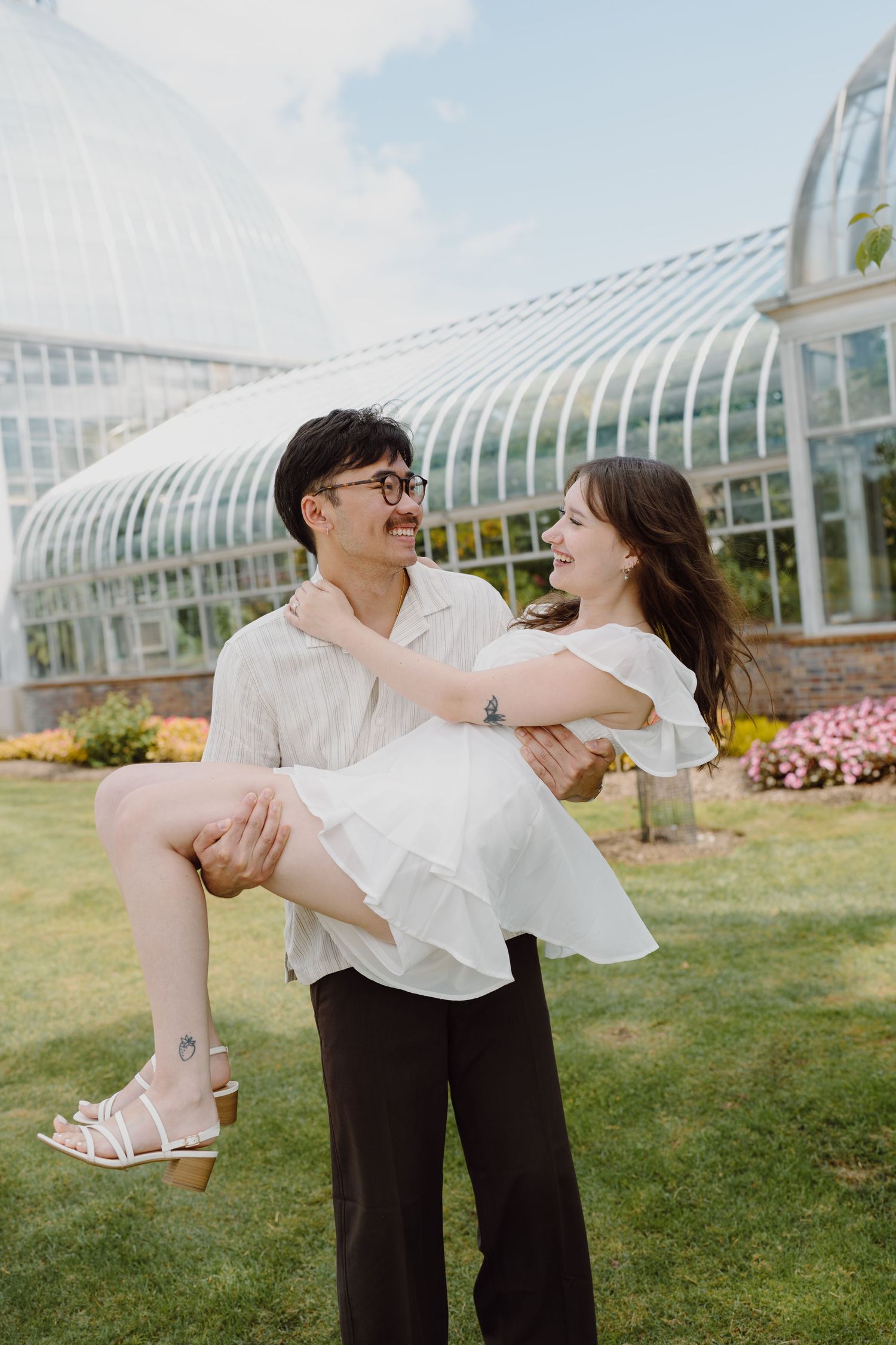 A couple dances playfully on a lawn in front of a glass conservatory building on a sunny day.