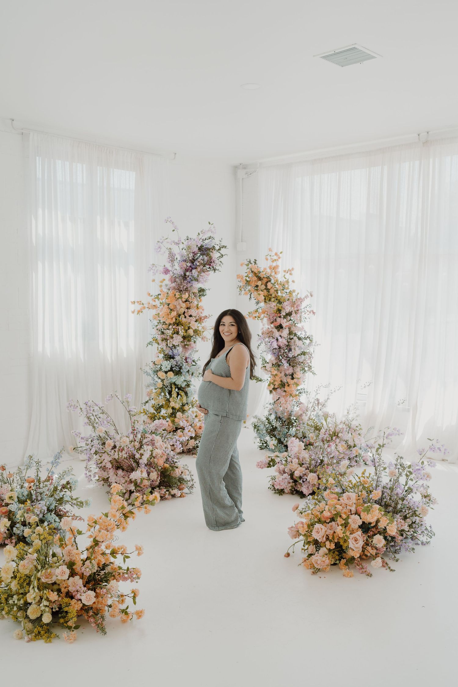 Side view maternity portrait with pastel floral arrangements in bright white studio setting.