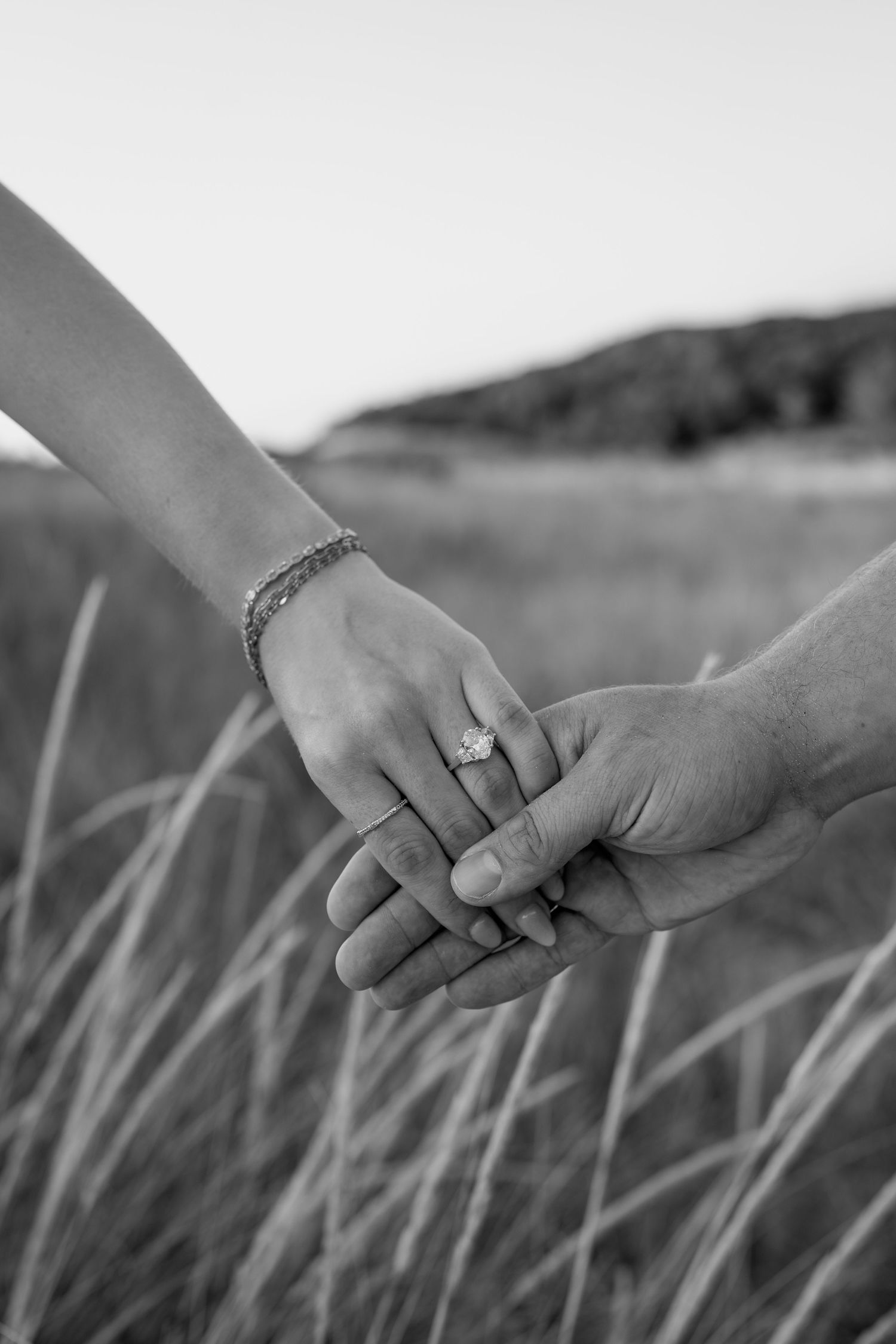 Black and white photo of hands with engagement rings touching against beach grass.