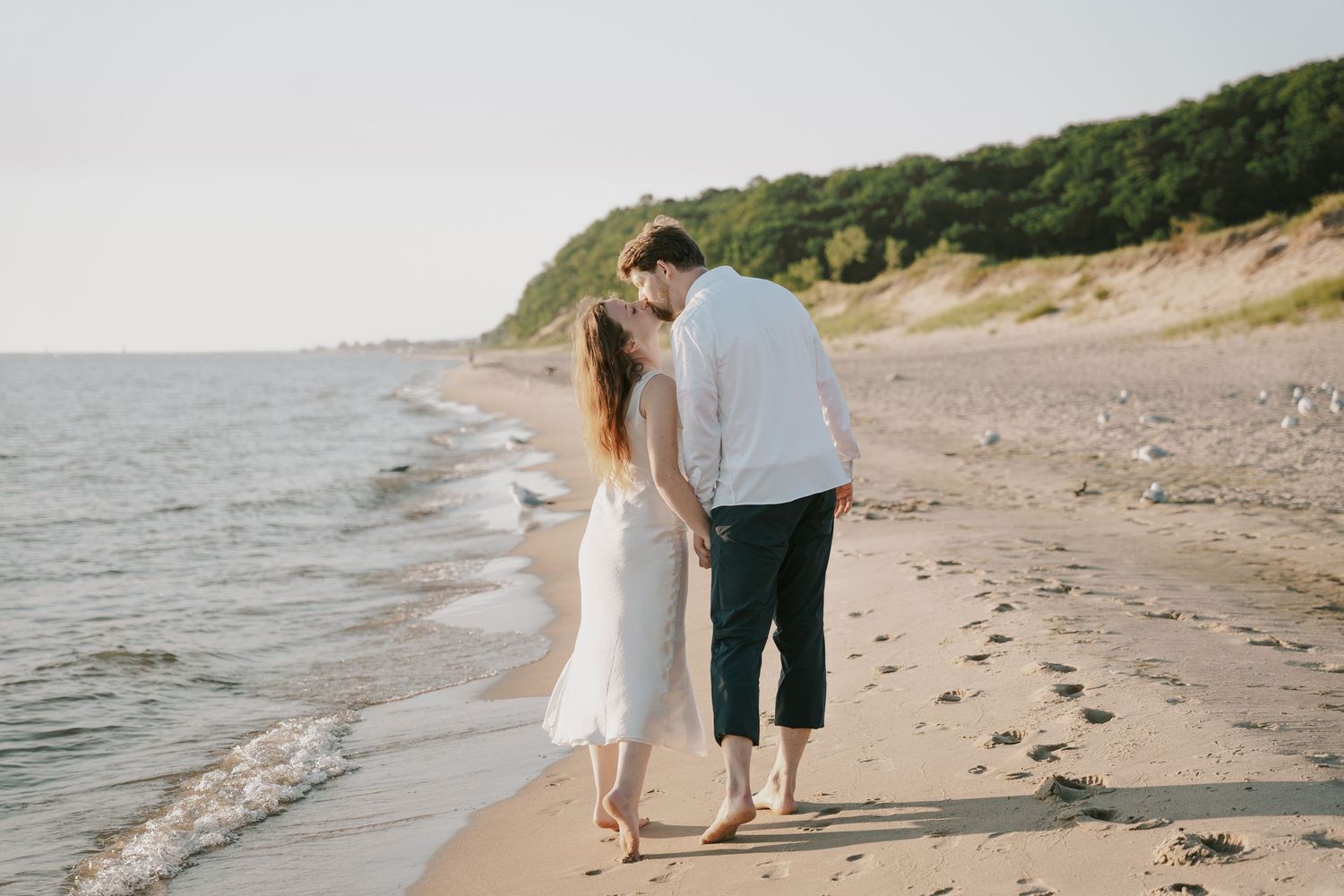 A couple walks hand in hand along a scenic beach shoreline with cliffs in the background.