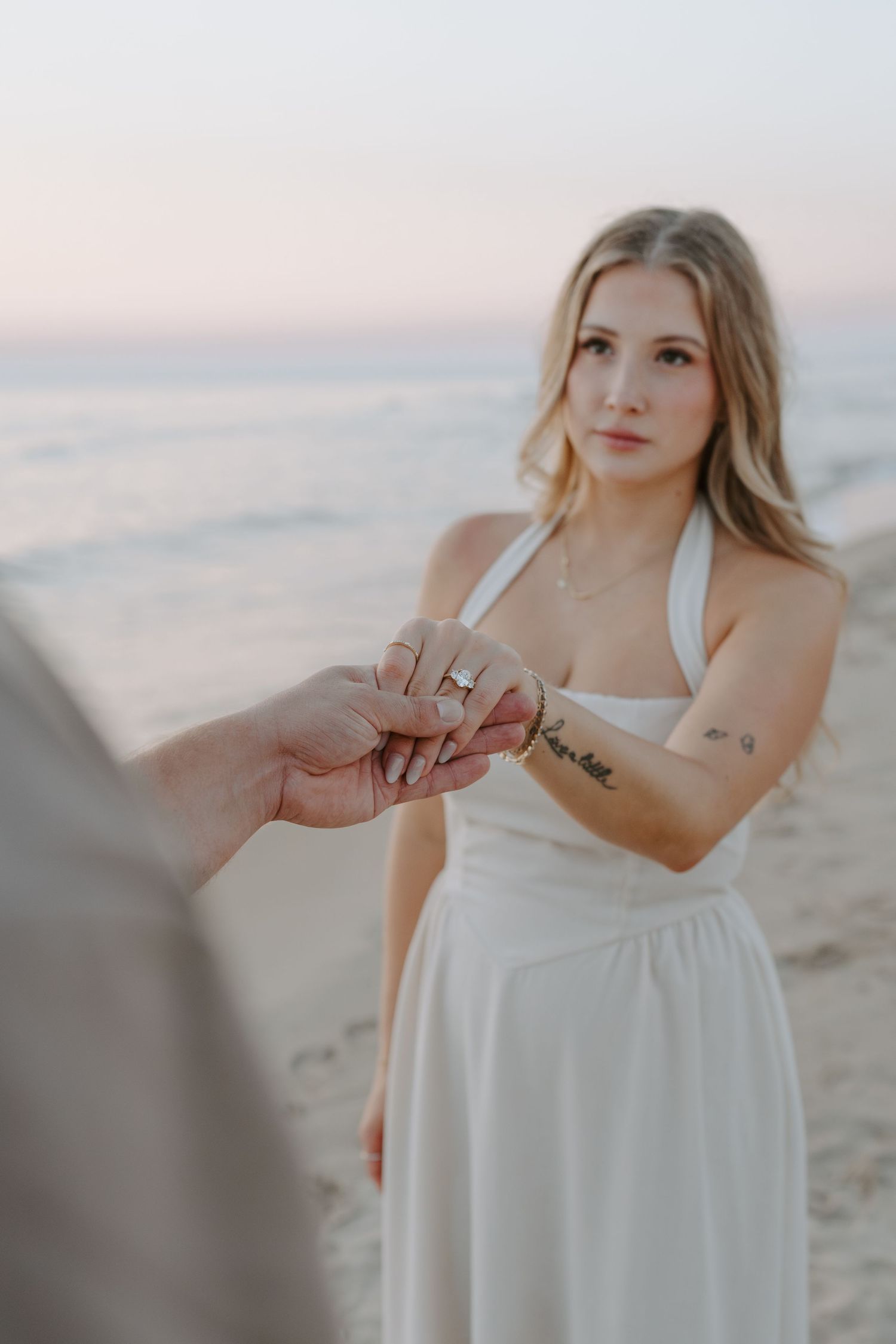 Woman in white dress showing engagement ring during sunset beach photoshoot.
