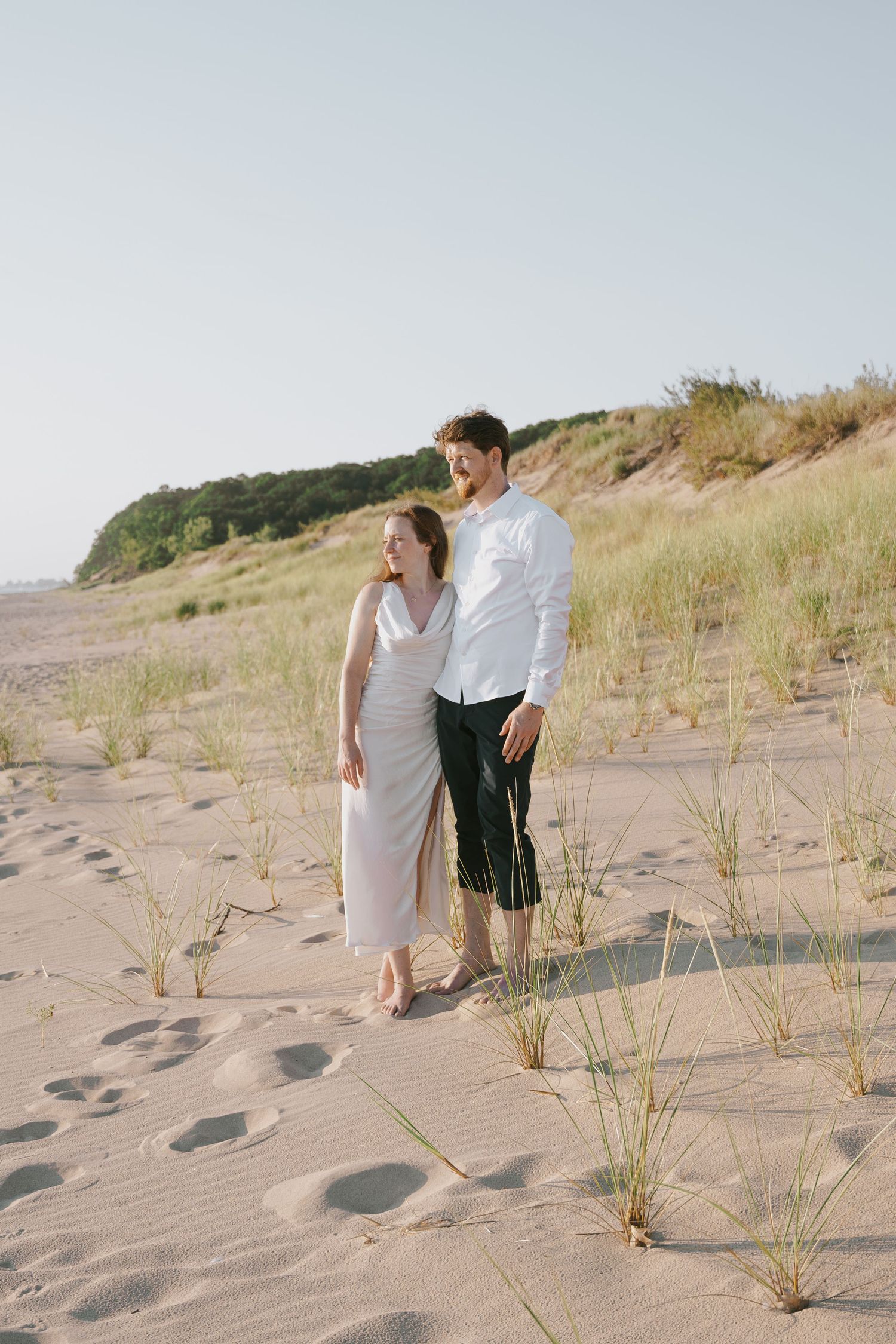 Two people stand together on coastal sand dunes with beach grass swaying in the breeze.