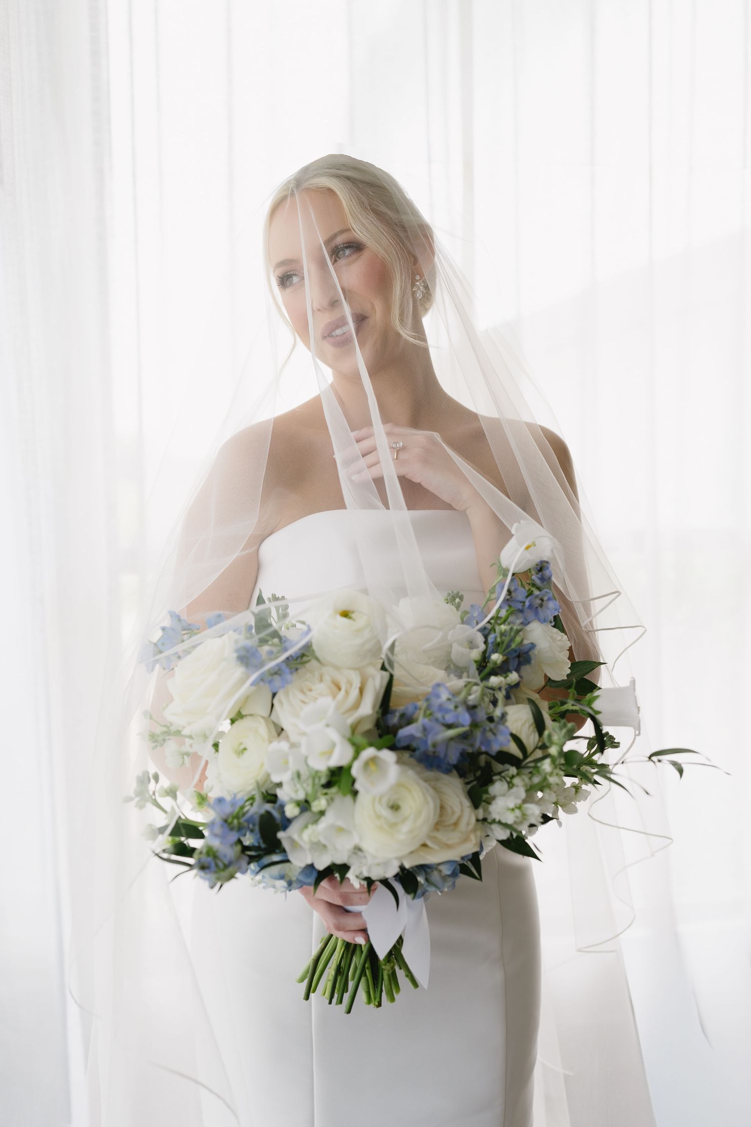 Bride in white strapless gown holding blue and white floral bouquet, draped in veil near sheer curtains.
