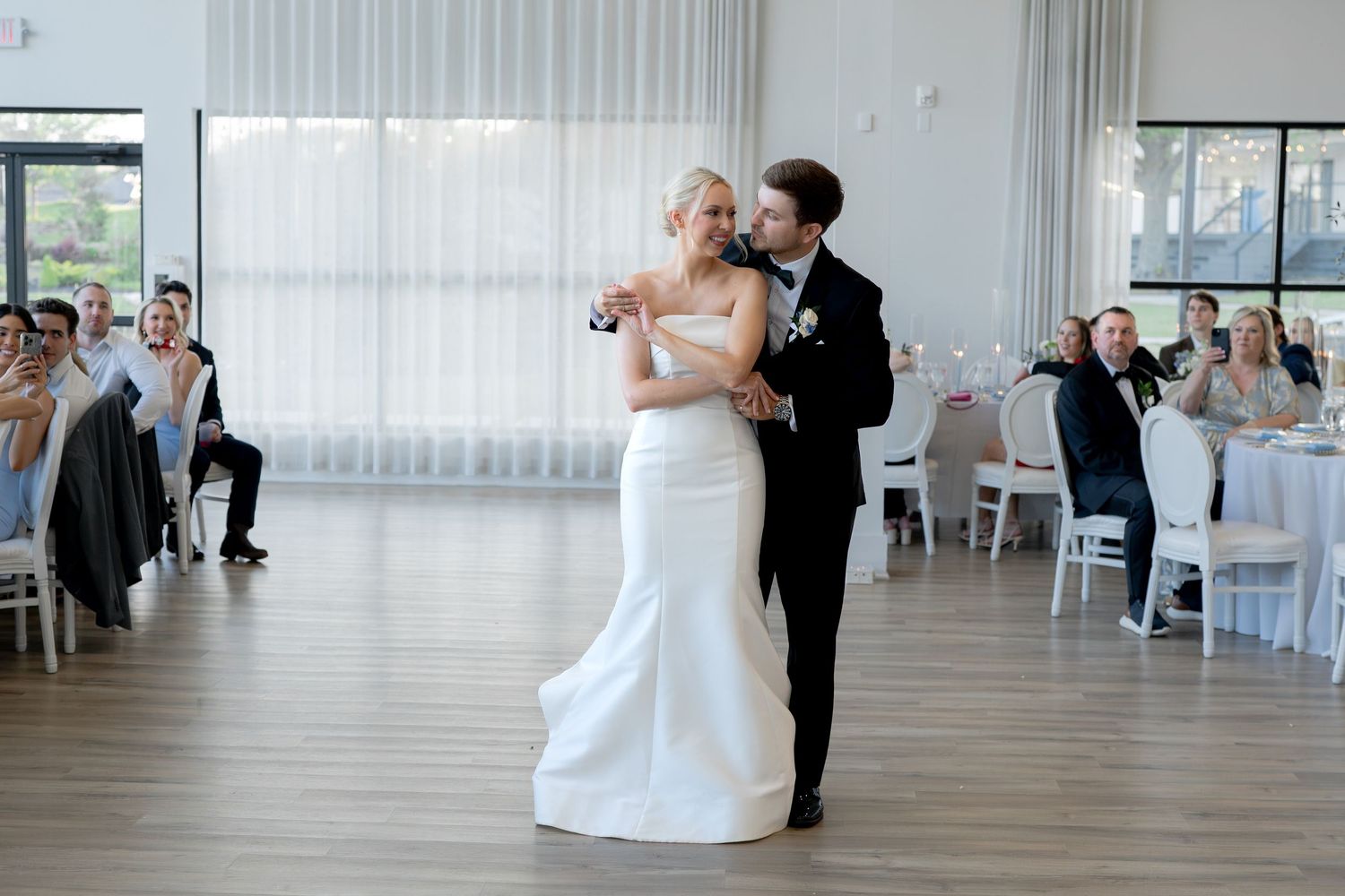 Bride and groom share their first dance in an elegant white wedding reception hall with guests watching.