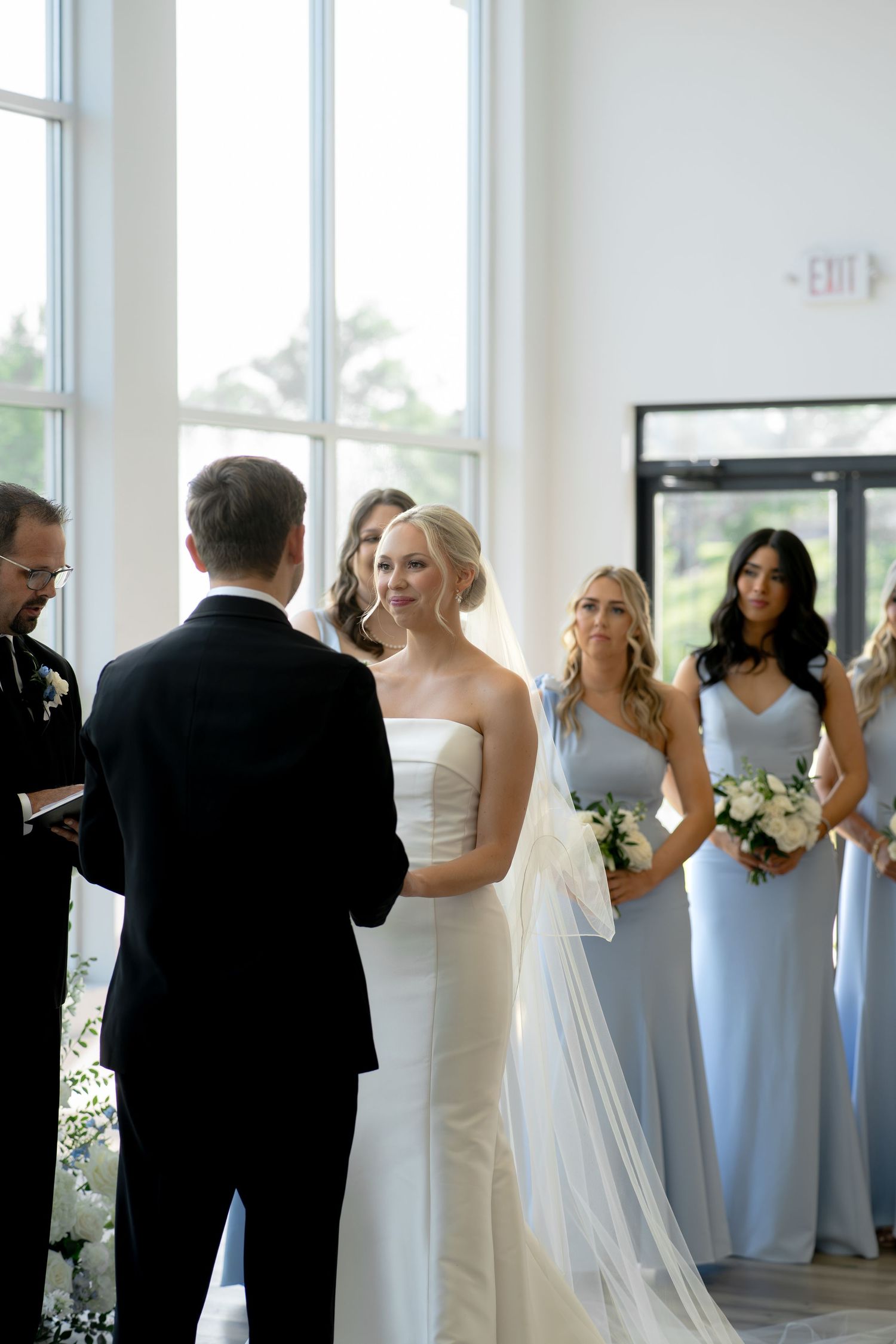 Bride in strapless white gown exchanges vows with groom as bridesmaids in blue dresses look on during indoor ceremony.