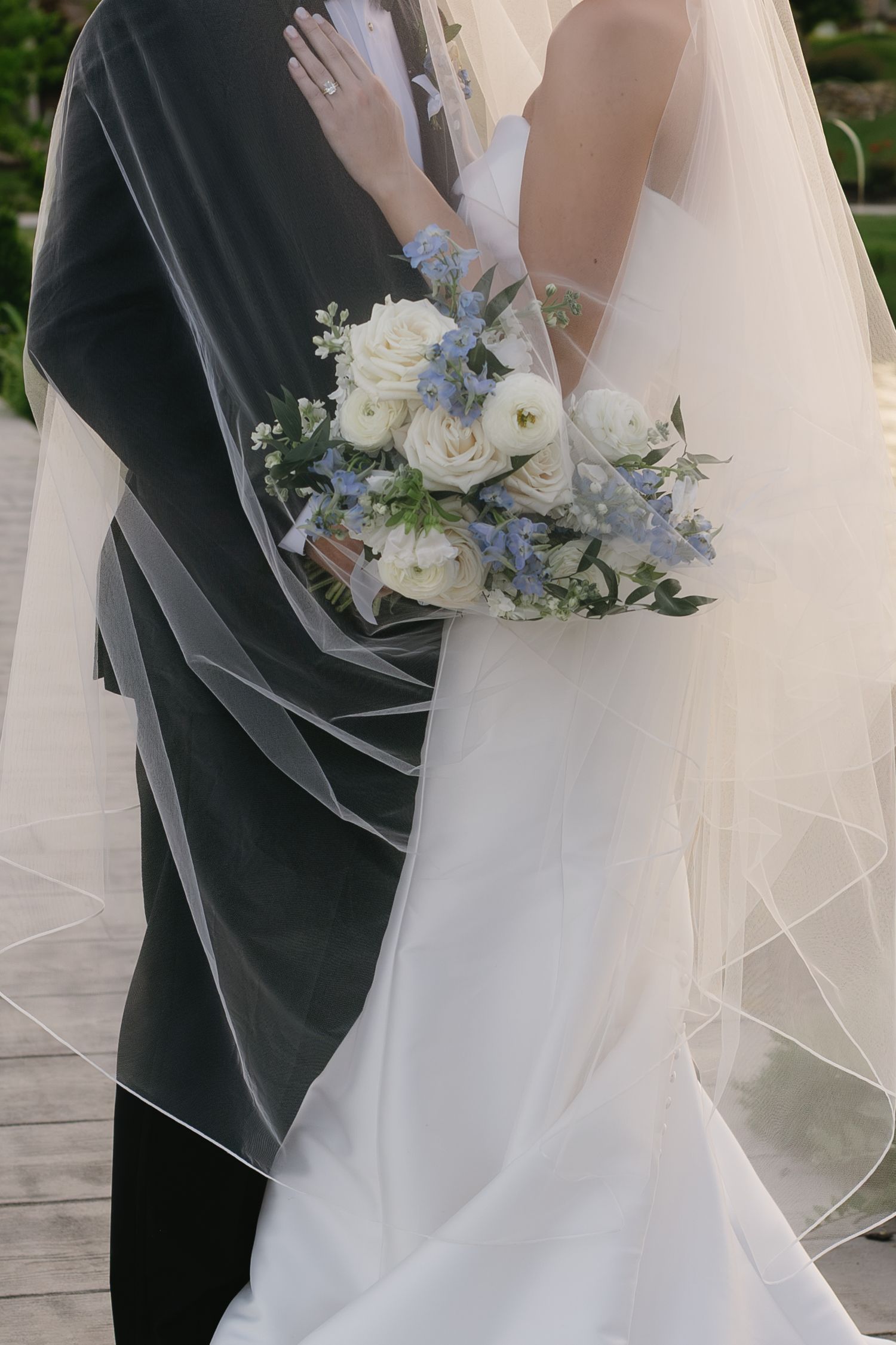 Bride and groom embracing, bride holding white and blue floral bouquet, wedding veil draped elegantly around couple.