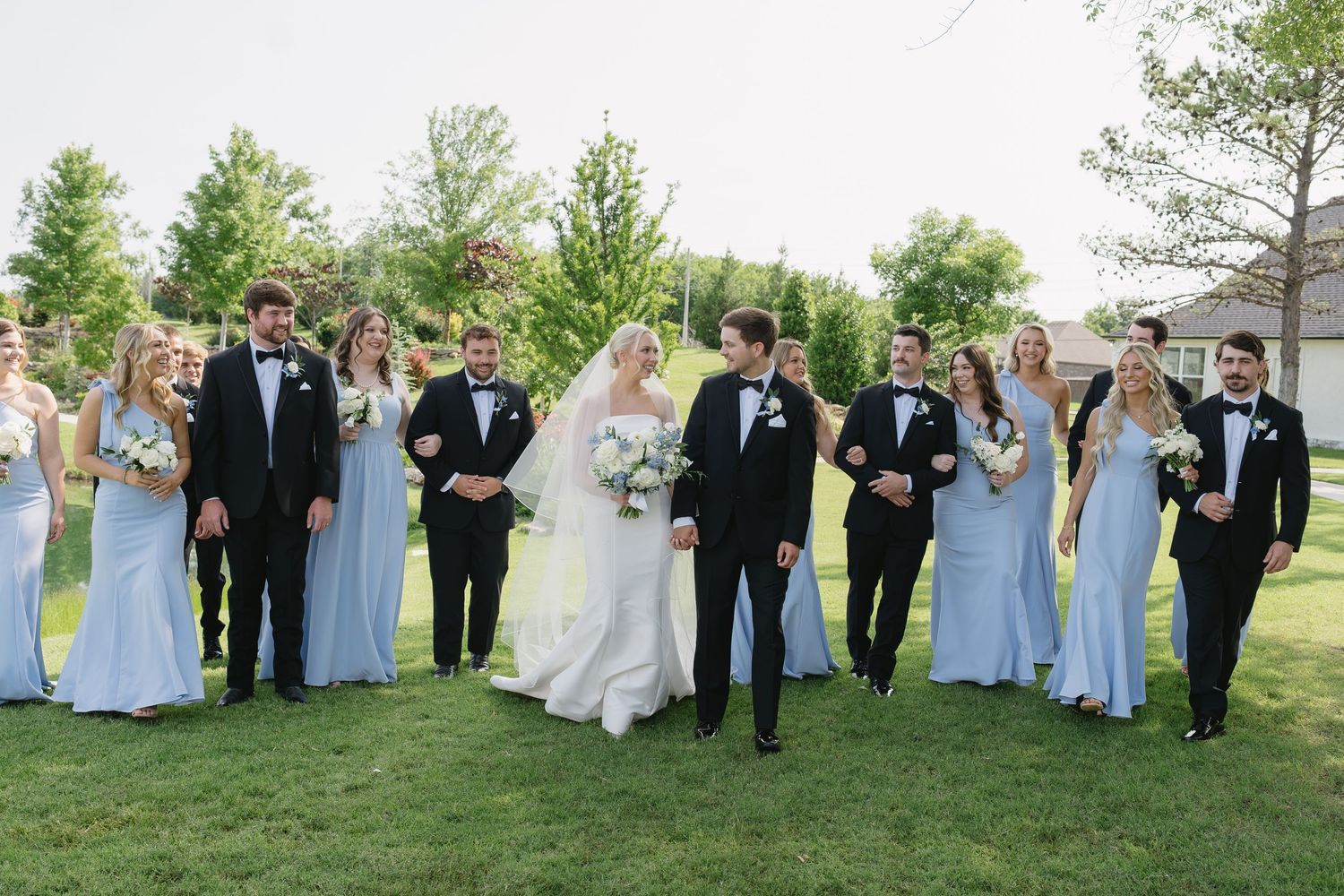 Wedding party walks outdoors on green lawn, bride and groom centered, bridesmaids in light blue gowns, groomsmen in black tuxedos.