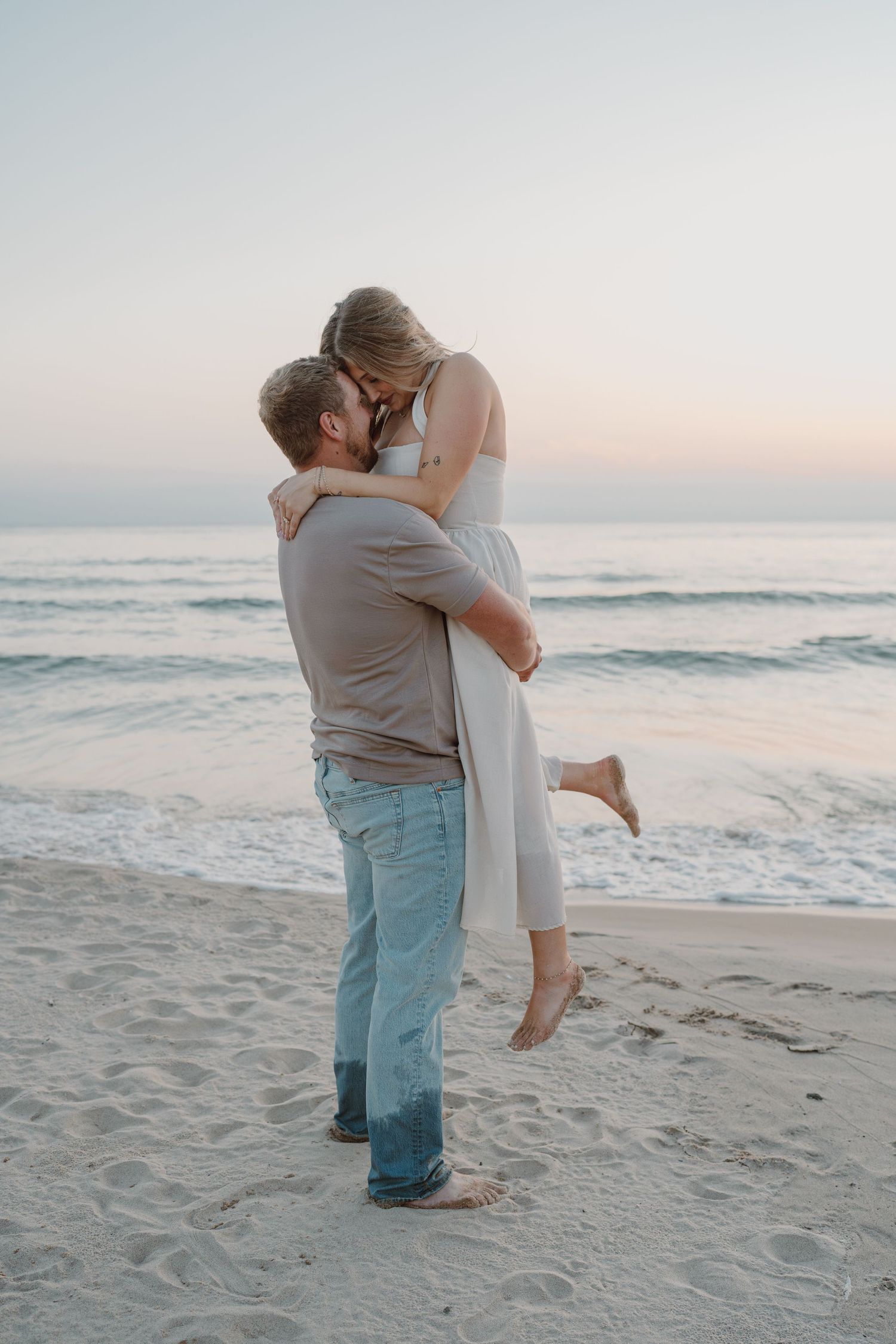 A couple embraces on a sandy beach during sunset with gentle waves in the background.