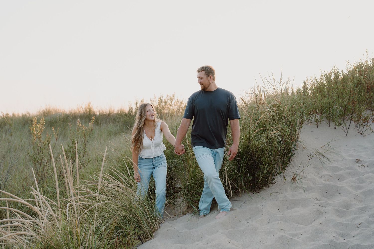 Two people hold hands while walking through coastal dunes with beach grass at golden hour.