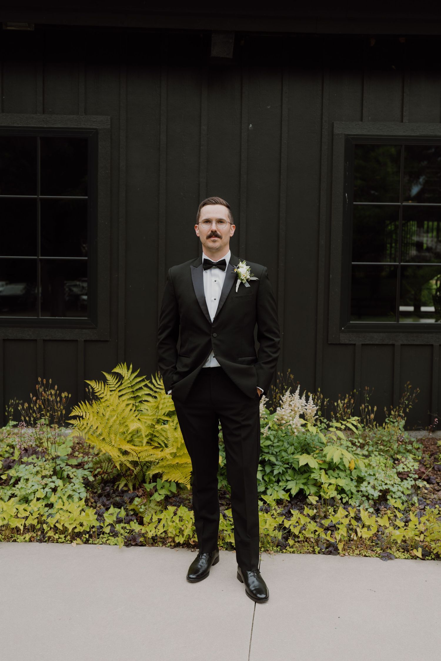 A person in a formal black tuxedo stands against a dark wall with yellow ferns in the garden.