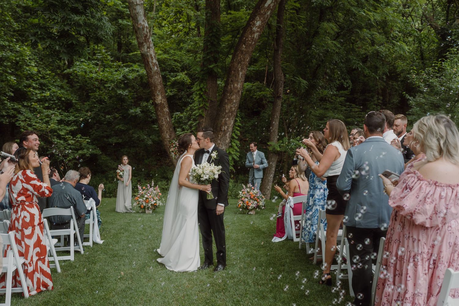 Guests seated in white chairs for an outdoor forest wedding with floral decorations.