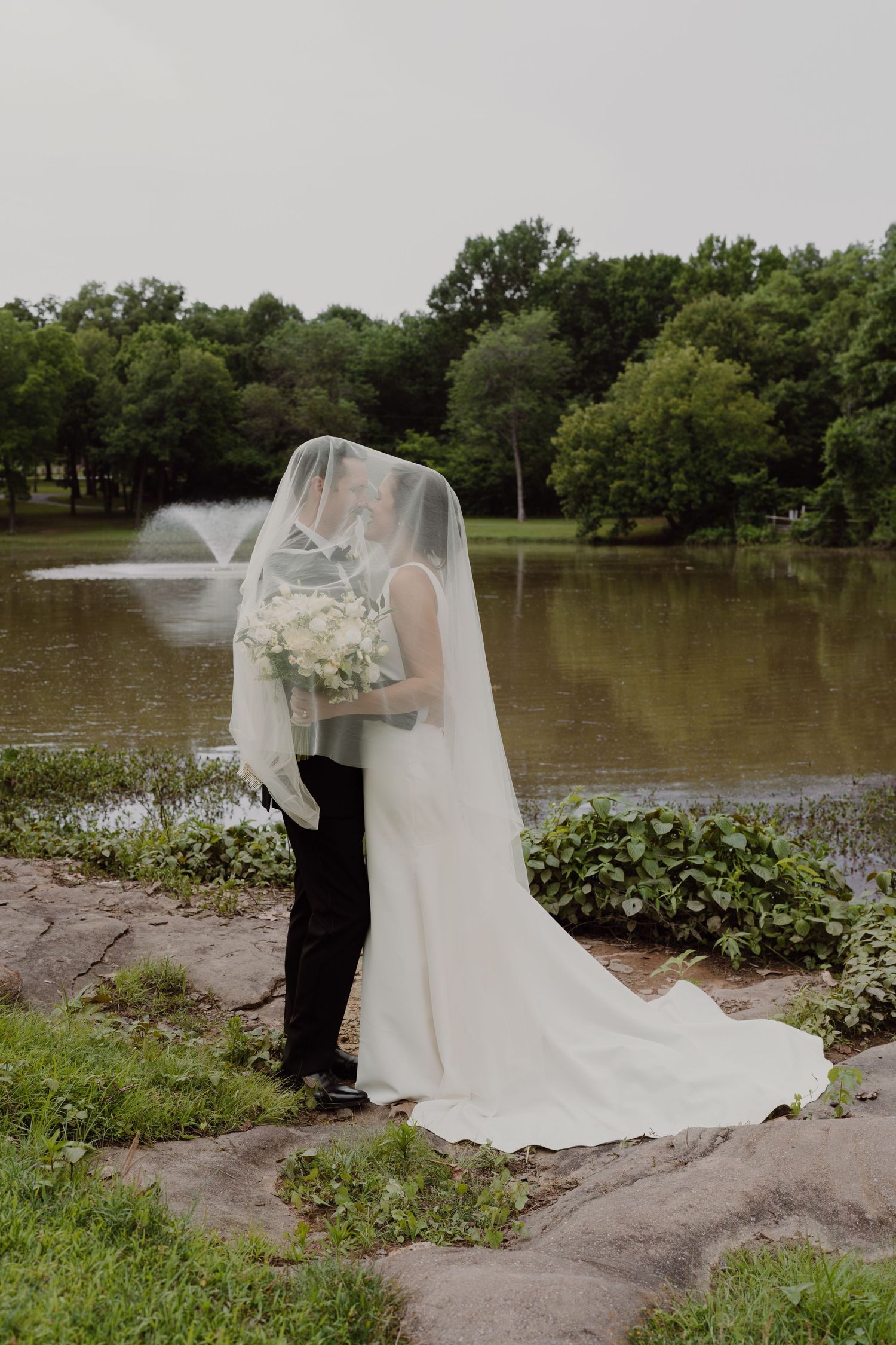 Wedding couple by a lake with flowing white dress and veil against natural landscape.