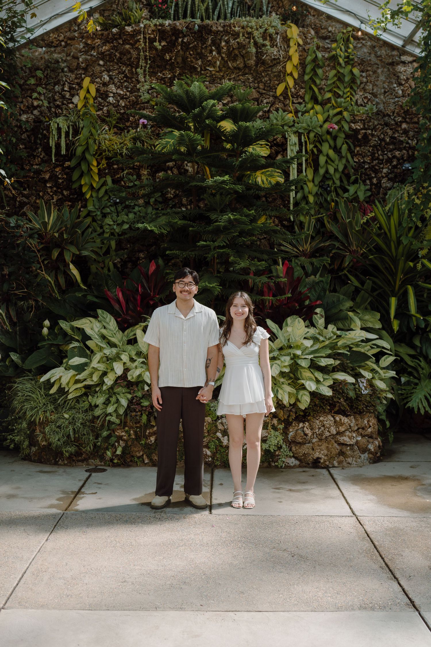 A couple walking together on a concrete path surrounded by tropical plants in a greenhouse.