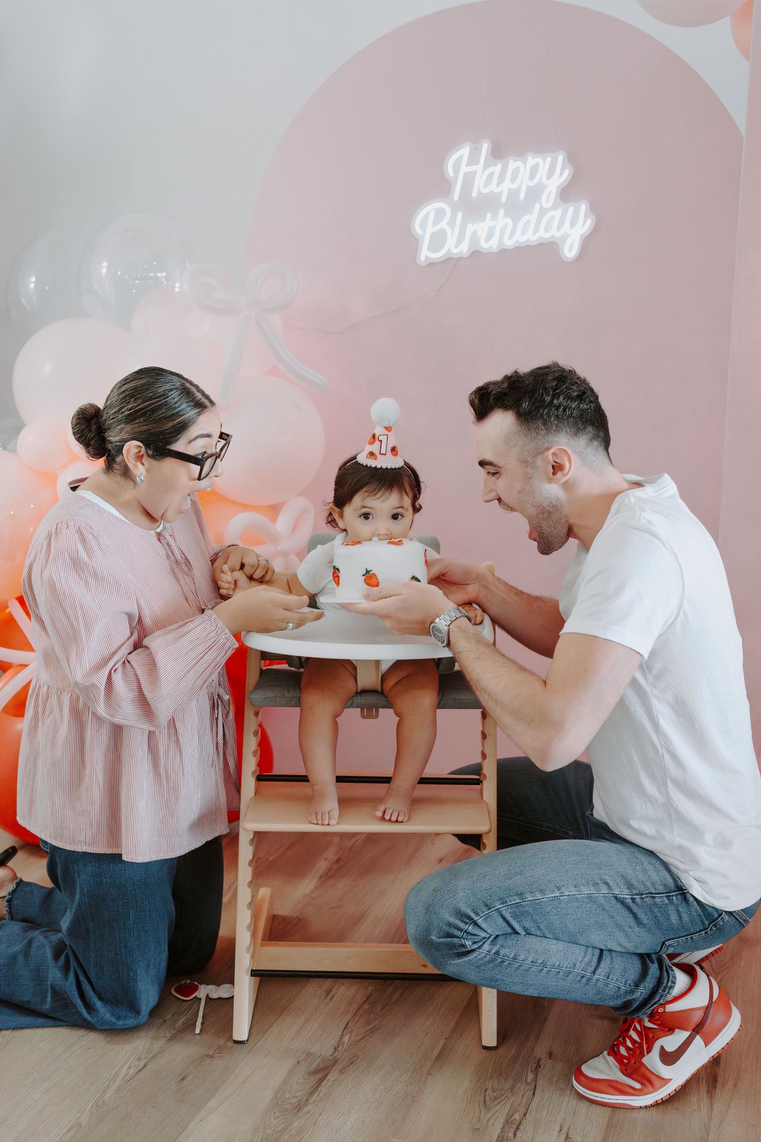 Birthday celebration scene with baby in wooden high chair and pink party decorations.