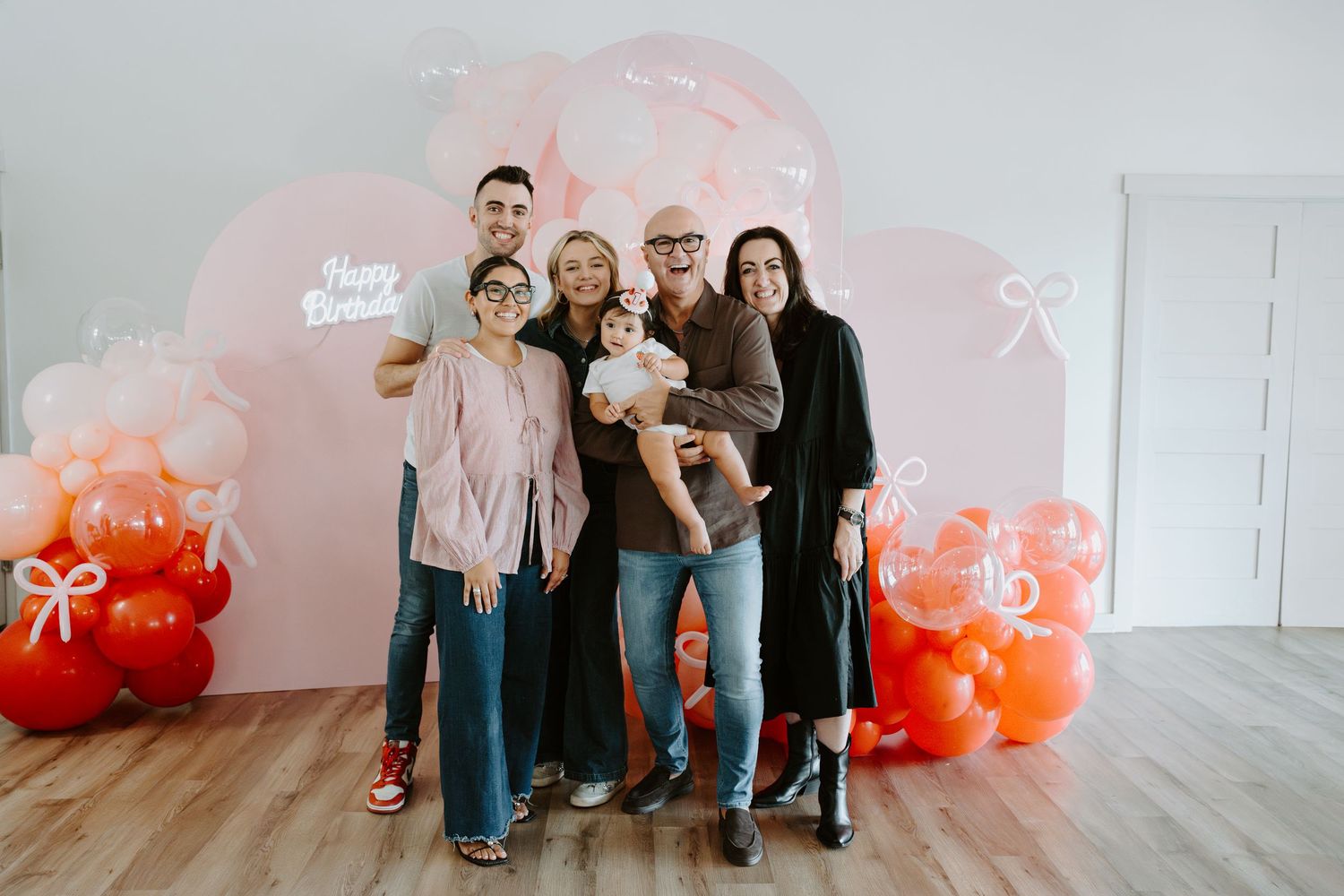 A group portrait in front of a pink balloon arch with 'Happy Birthday' text visible.