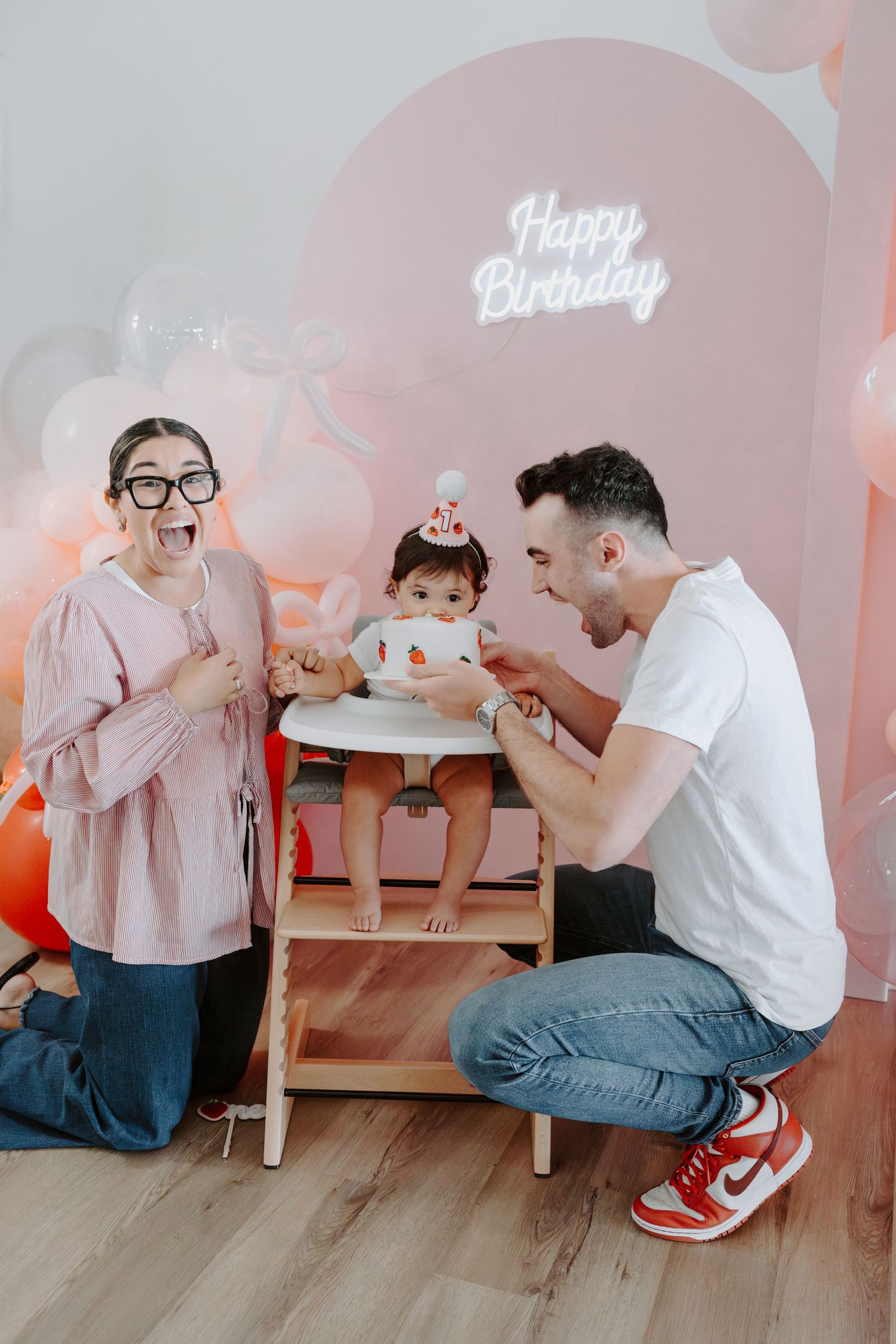 Birthday photoshoot scene with pink balloons and neon sign showing candid family moments around a small table.