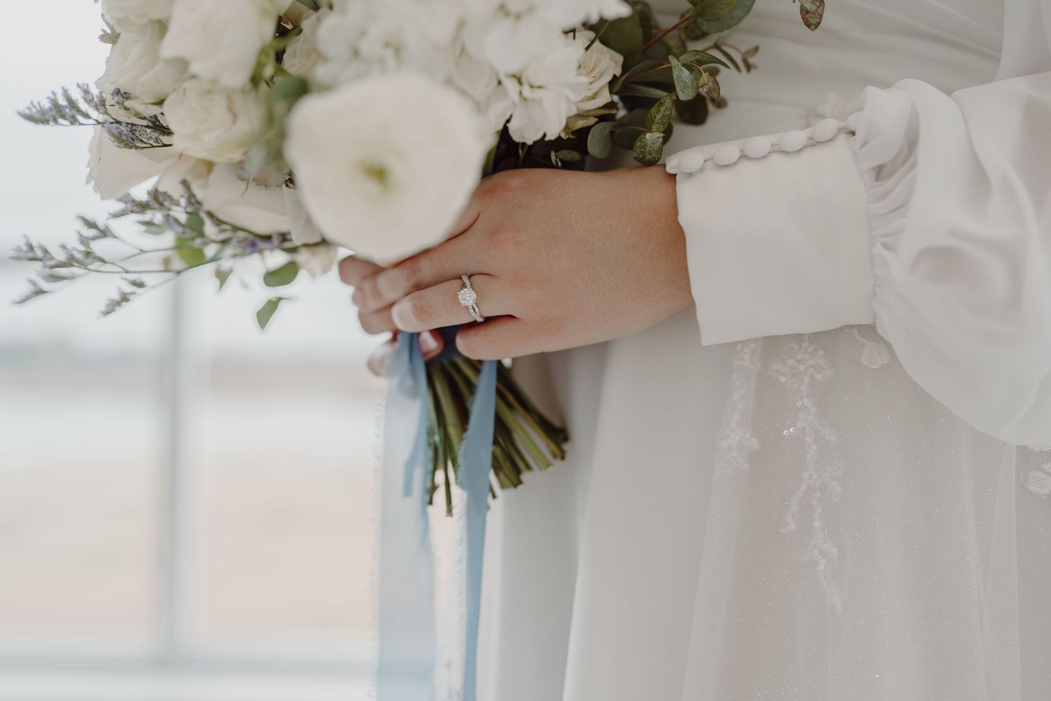 Close-up of hands holding white wedding bouquet with engagement ring visible.