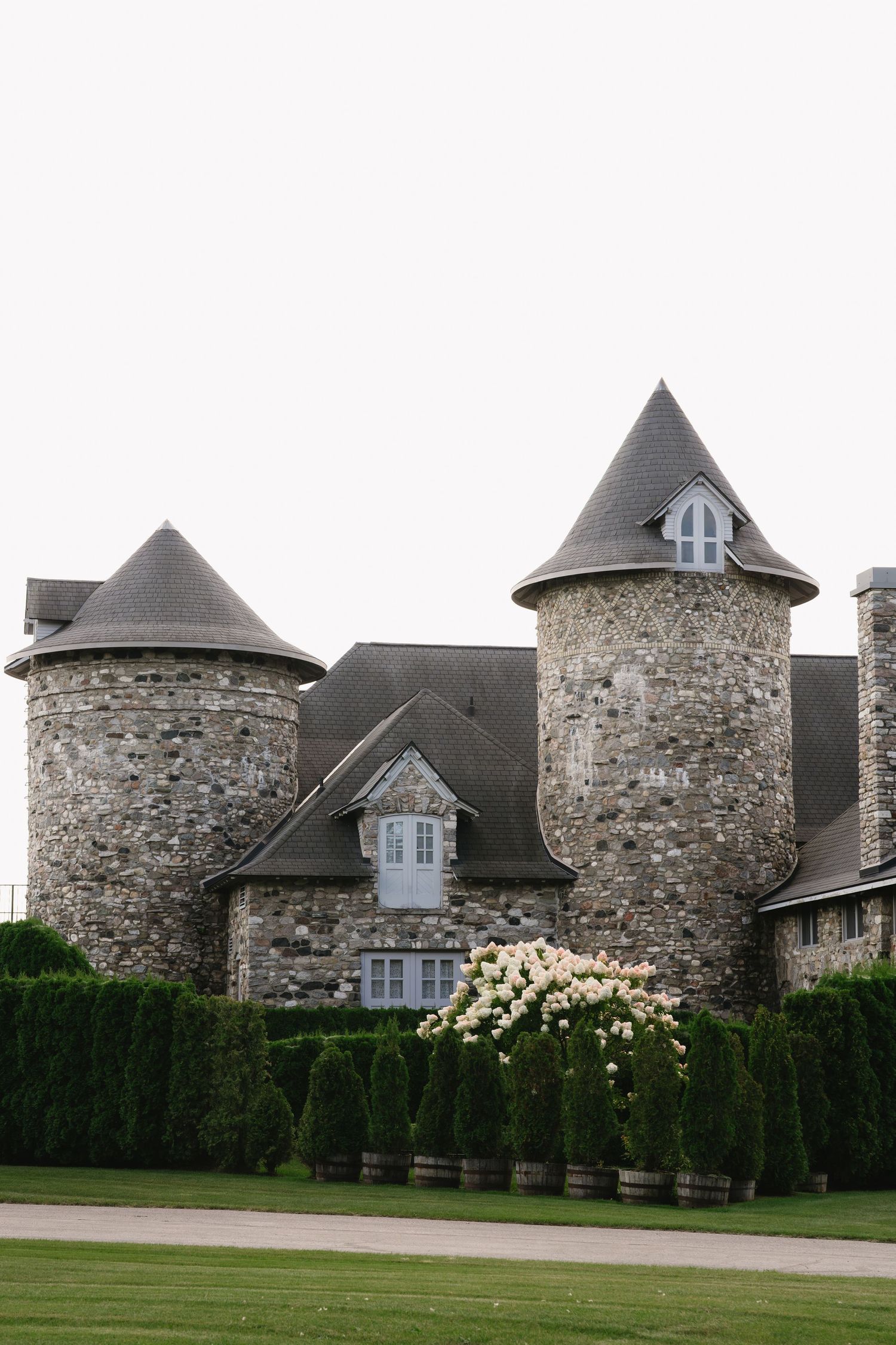 Stone castle-style building with twin turrets, slate roofs, and manicured hedges with white flowering shrubs.