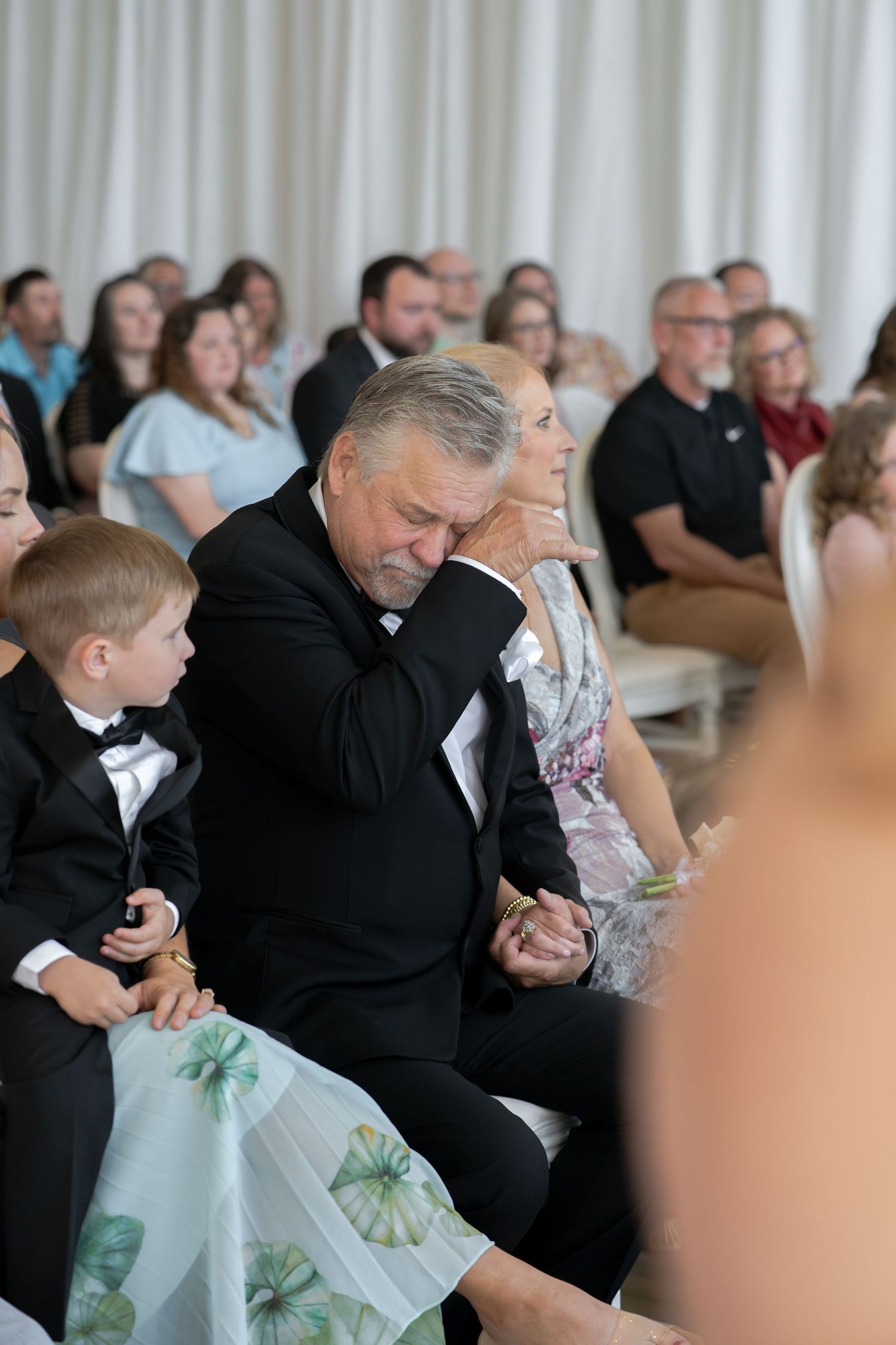 Elderly man in black suit bowing head emotionally during a wedding ceremony while seated among guests.
