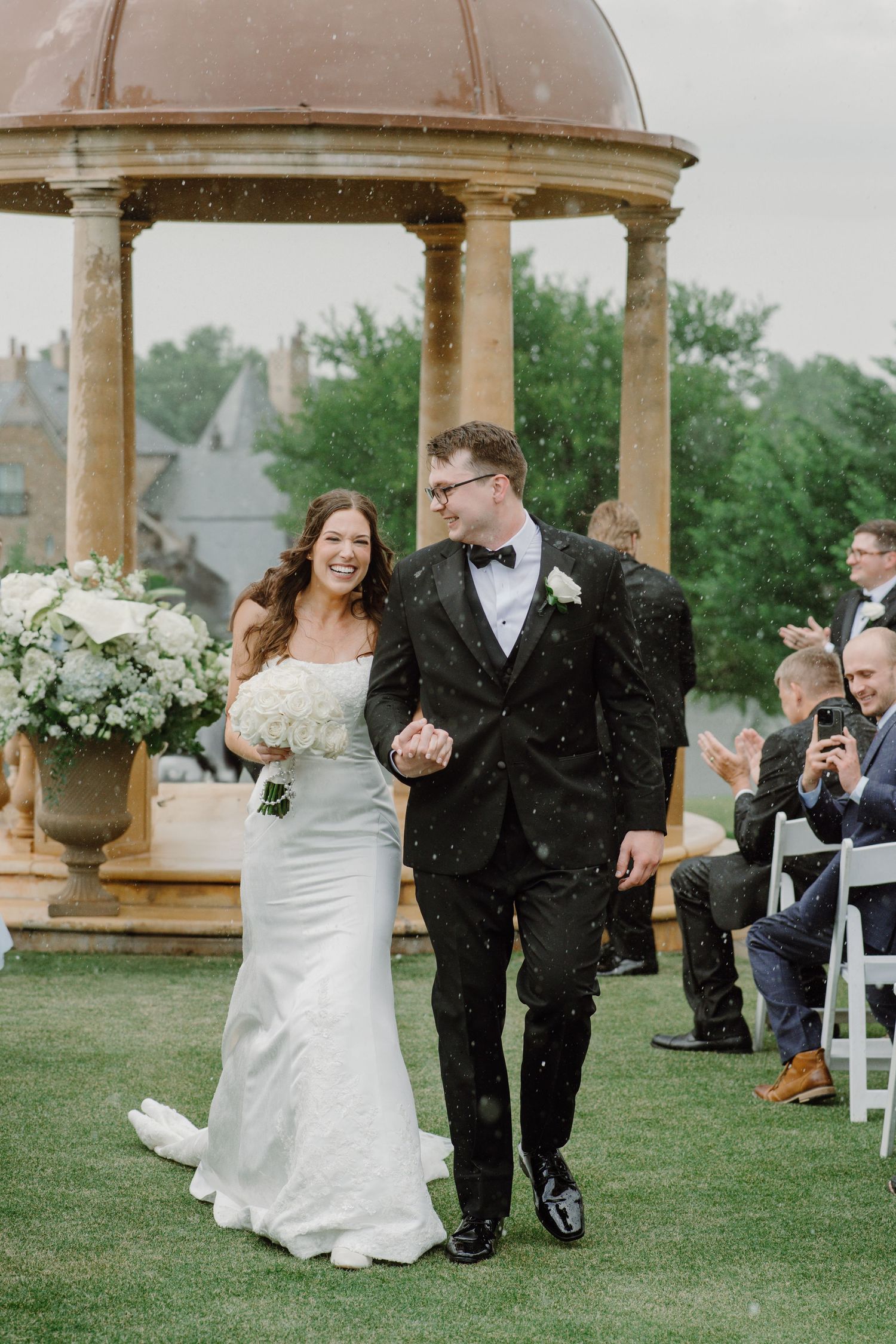 A couple laughs and runs through falling rain during their outdoor wedding ceremony under a stone gazebo.