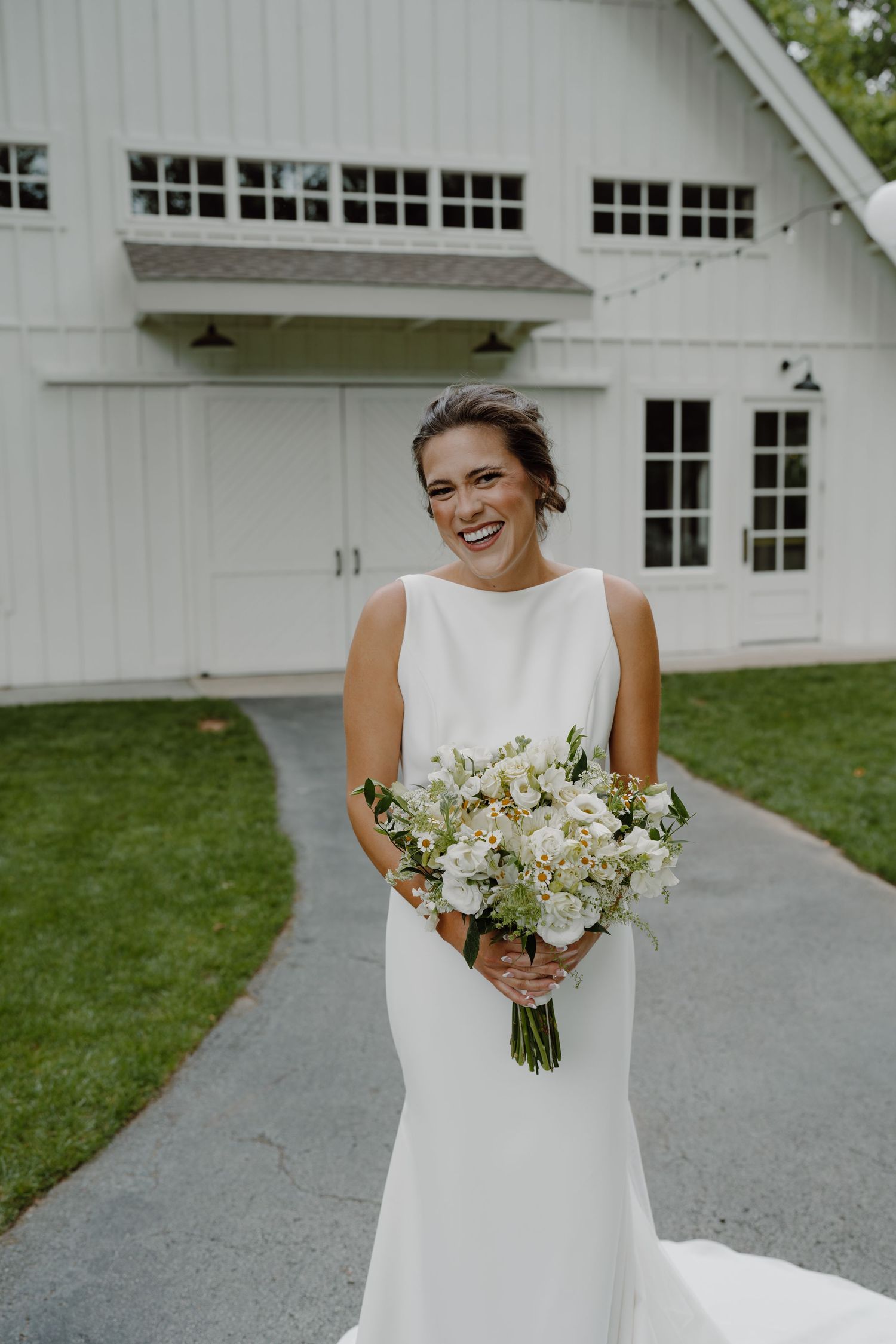 A bride holds a lush white bouquet while posing near a white barn building with string lights.